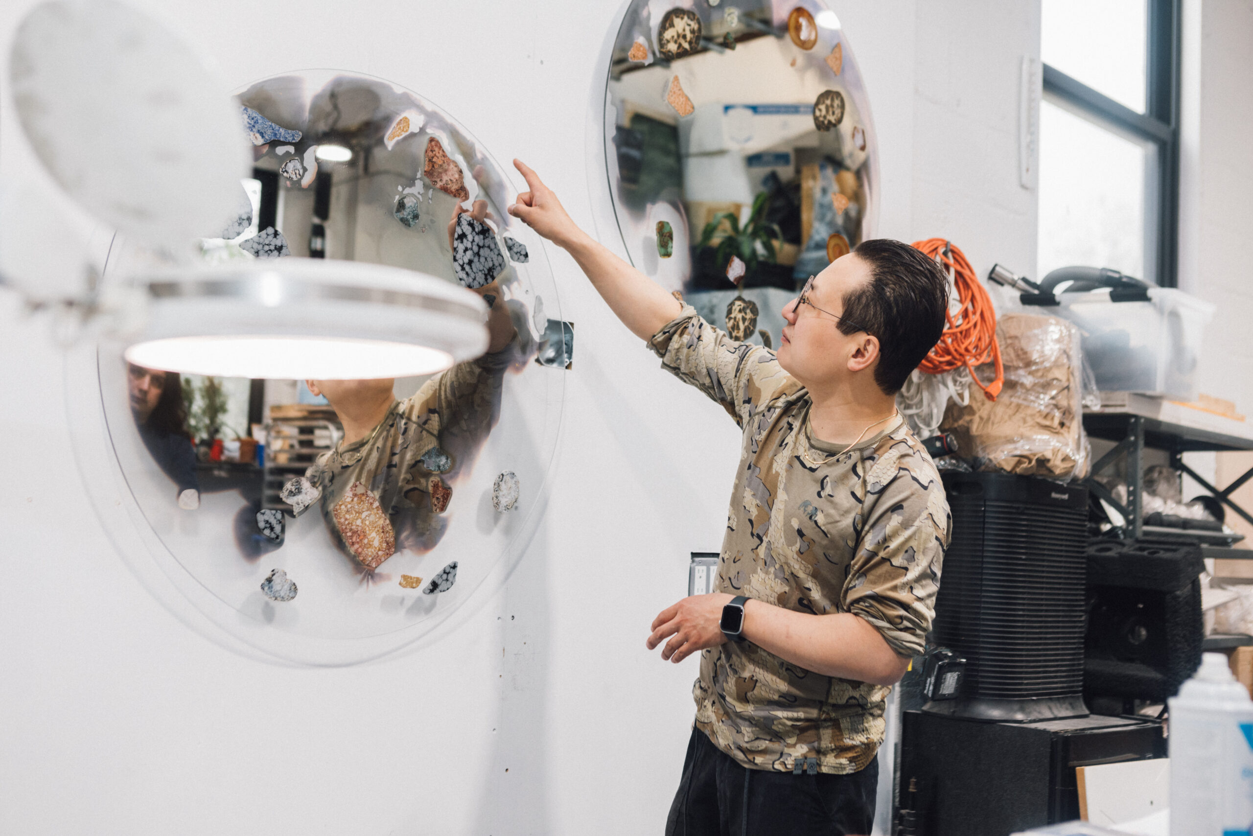 A person in a patterned shirt is pointing at a circular mirror mounted on a wall, which features various colorful stones embedded in it. The setting is a workshop with shelves holding supplies and equipment. A bright light is visible in front of the person, reflecting some of their features in the mirror.