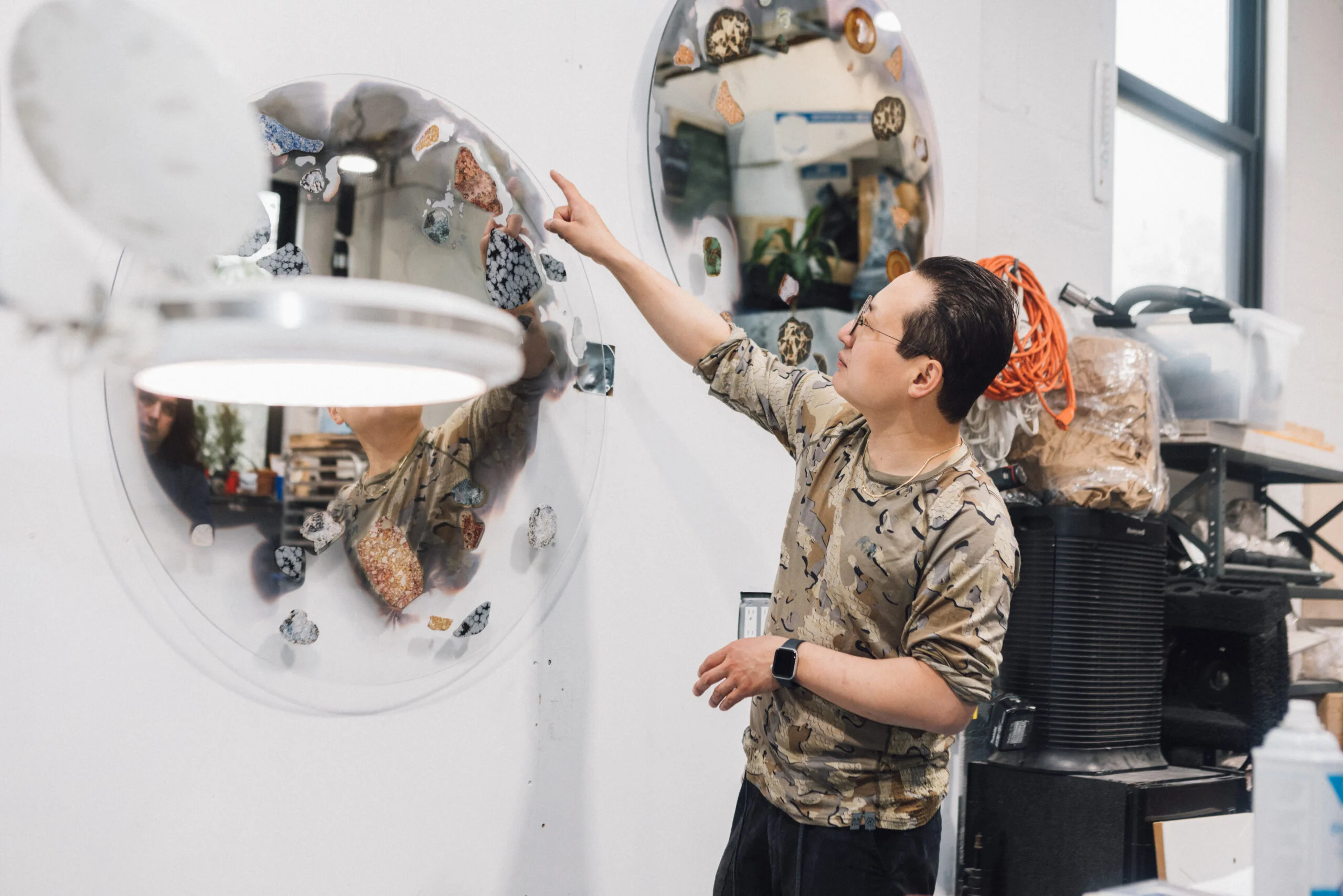 A person in a patterned shirt is pointing at a circular mirror mounted on a wall, which features various colorful stones embedded in it. The setting is a workshop with shelves holding supplies and equipment. A bright light is visible in front of the person, reflecting some of their features in the mirror.
