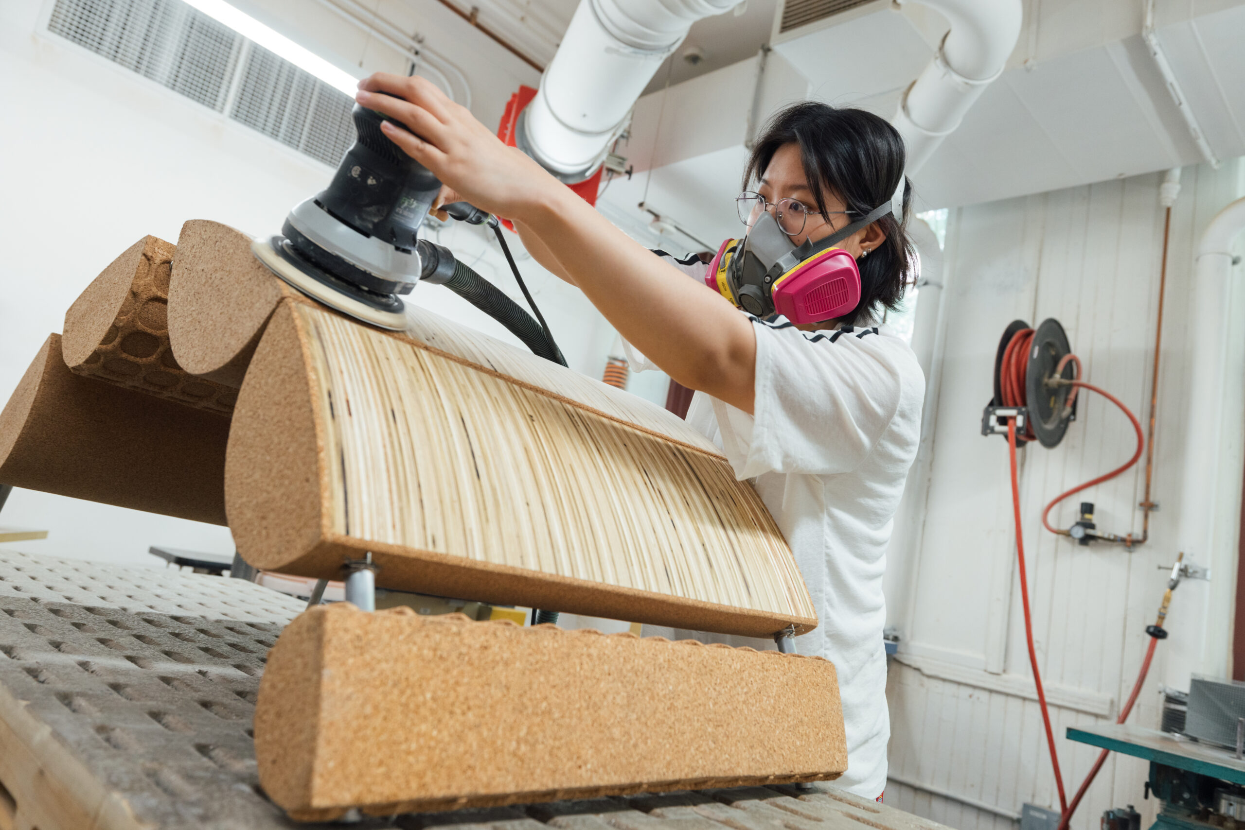 Student wearing safety goggles and a respirator mask uses a handheld orbital sander to smooth a curved laminated wood panel secured on a raised sanding table inside a woodshop, with ventilation ducts, tools, and work surfaces visible in the background.