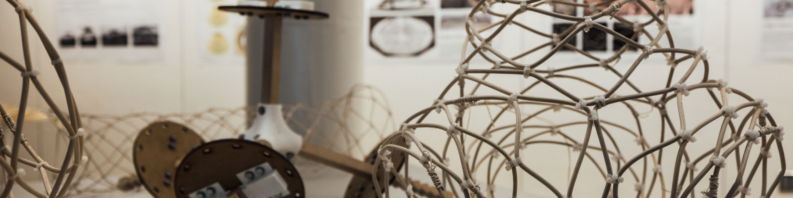 An art installation featuring intricate, lattice-like structures made from thin rods and ropes. Various geometric forms are scattered throughout the space, with a background displaying informational posters. Some structures rest on wooden disks, suggesting an exploratory or experimental theme in the design. The overall atmosphere appears artistic and modern.