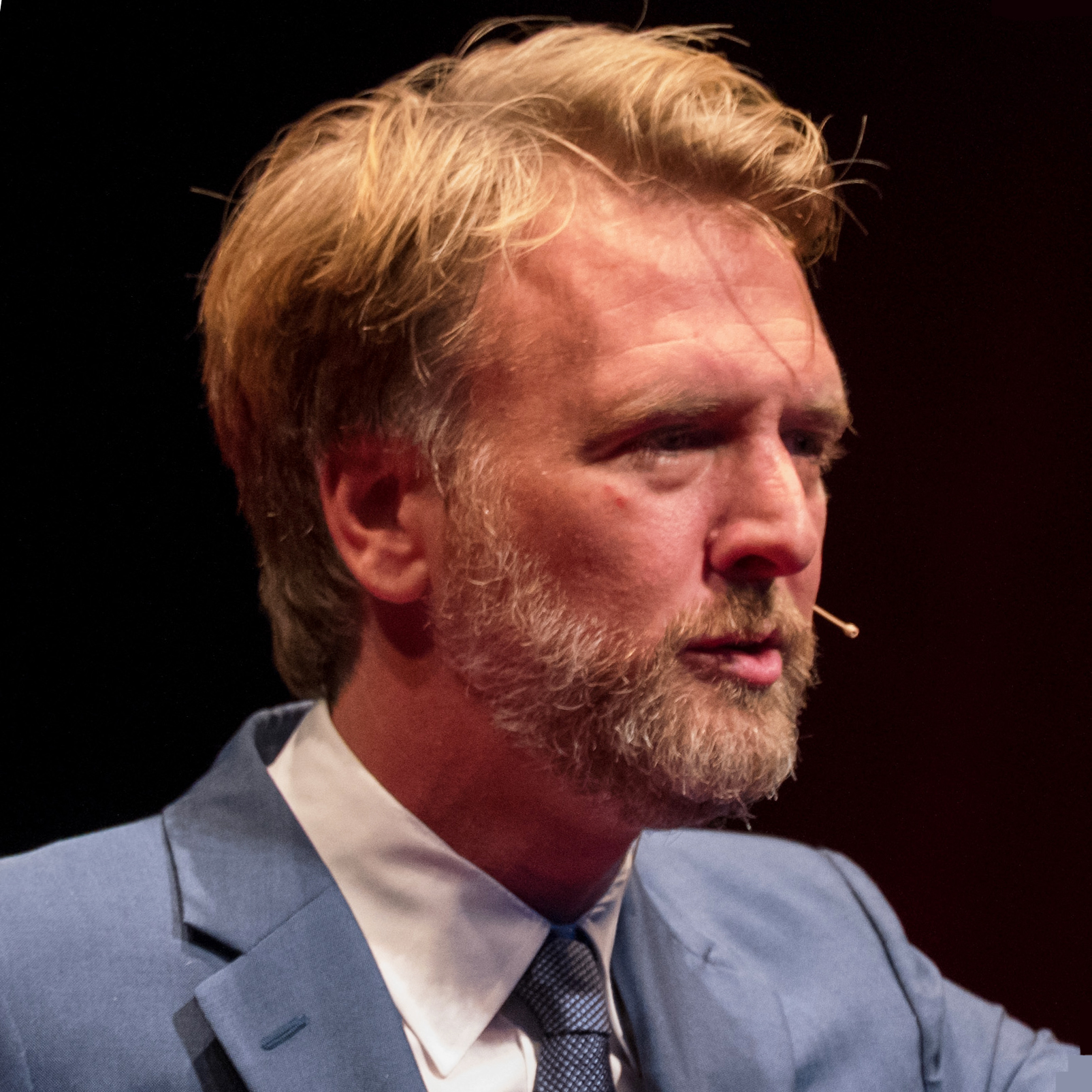 A man with a well-groomed beard and tousled light brown hair is speaking on stage. He is wearing a blue suit and white shirt, and has a small microphone attached to his cheek. The background appears dark, emphasizing his expression and attire.