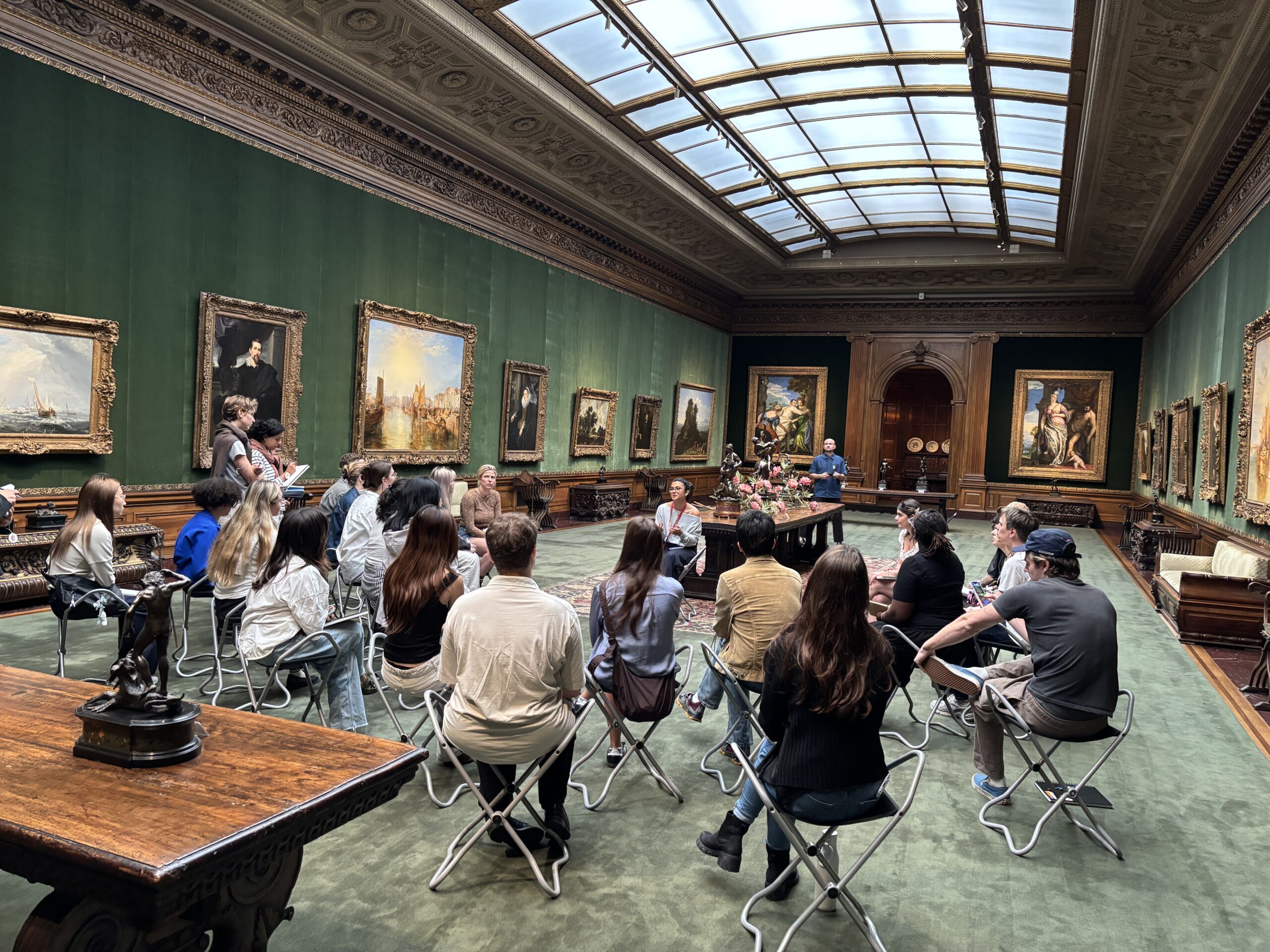 A group of around twenty people is seated in a spacious art gallery with green walls, surrounded by framed paintings. They sit in folding chairs, facing a presenter at the front, who appears to be speaking. In the background, several large paintings and a wooden display cabinet are visible, with natural light coming through a skylight above.