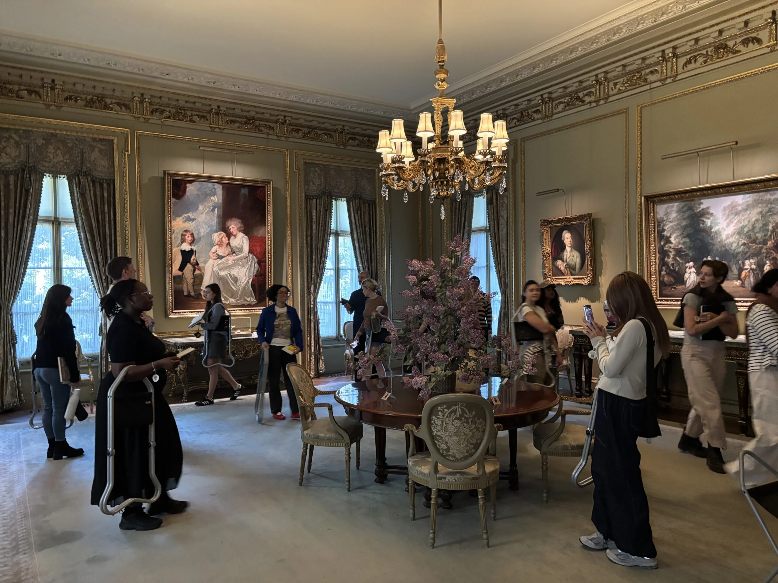 A spacious, elegantly decorated room with ornate detailing and large windows. Several individuals are walking around, some looking at artworks while others take photos. A table with a floral centerpiece is in the middle, surrounded by chairs. Two large paintings hang on the walls, depicting portraits and a landscape. The ambiance is bright and showcases a classic interior design.