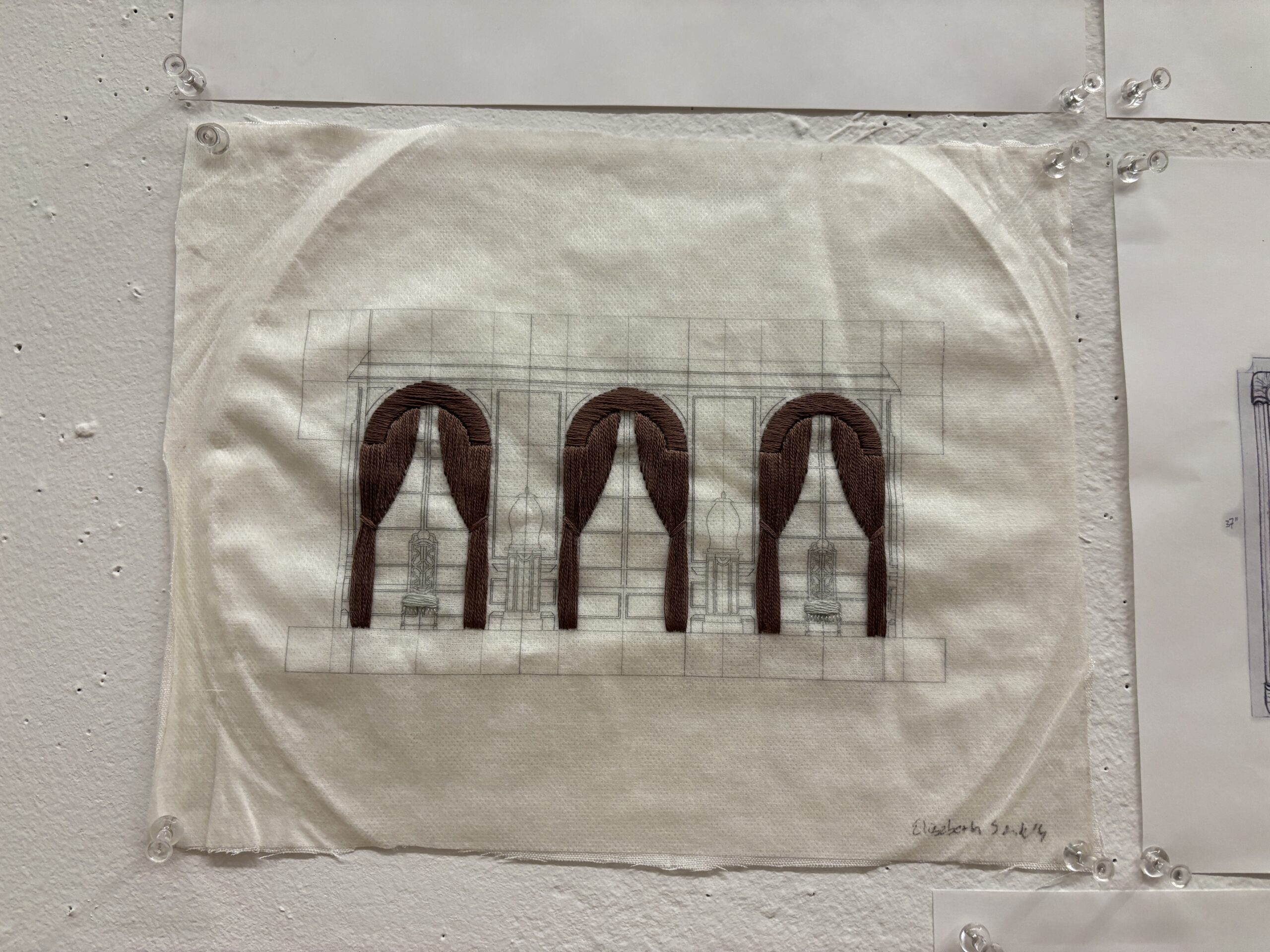 A textile artwork depicting three arched curtains in shades of brown. The curtains are embroidered on a light fabric, with faint outlines of architectural elements visible behind them. Below the curtains, there are stylized representations of objects, possibly vases or tables. The artwork is secured to a wall with small clips. The artist's name, 
