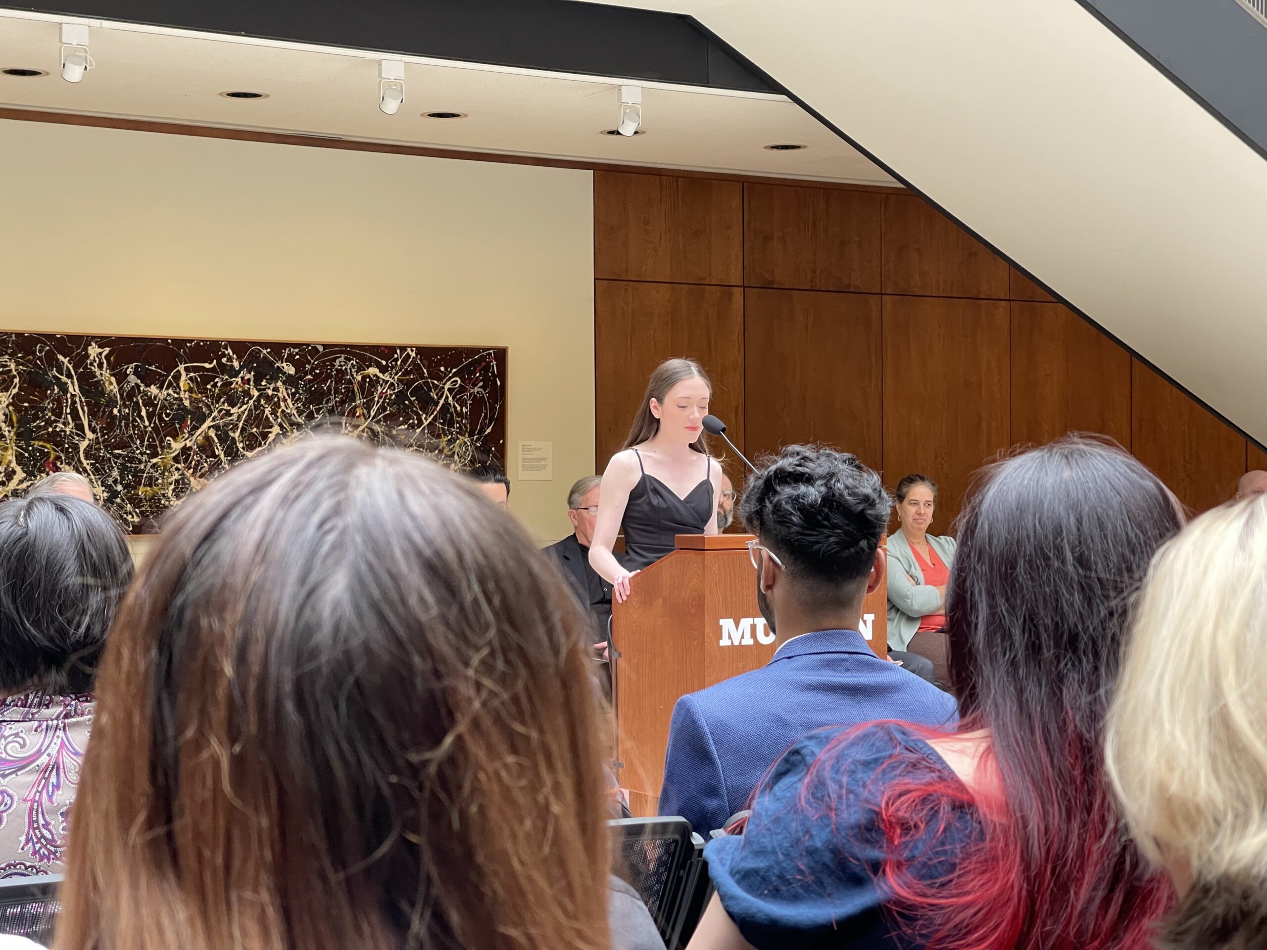 A young woman in a black dress stands at a podium speaking into a microphone, with a colorful abstract painting on the wall behind her. She is addressing an audience seated in front of her. Several people can be seen in the audience, including a man with curly hair and a woman in a teal jacket, all focused on her speech. The setting appears to be a formal event in a gallery or museum.