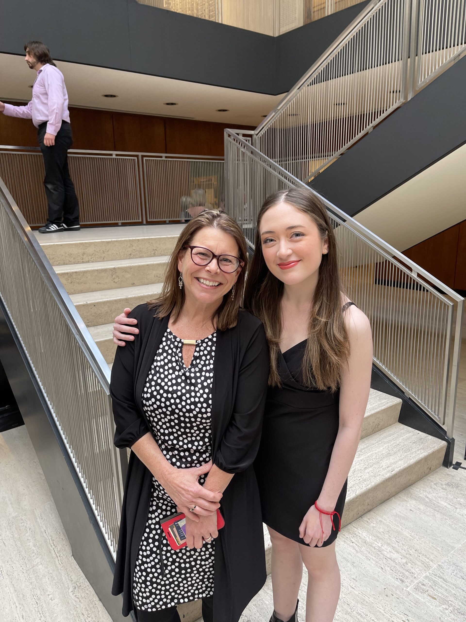 A young woman in a black dress speaks at a podium during an event, with a microphone in front of her. An abstract art piece hangs on the wall behind her, and several audience members are visible in the foreground, including people with diverse hairstyles. Attendees appear engaged, listening intently to the speaker.