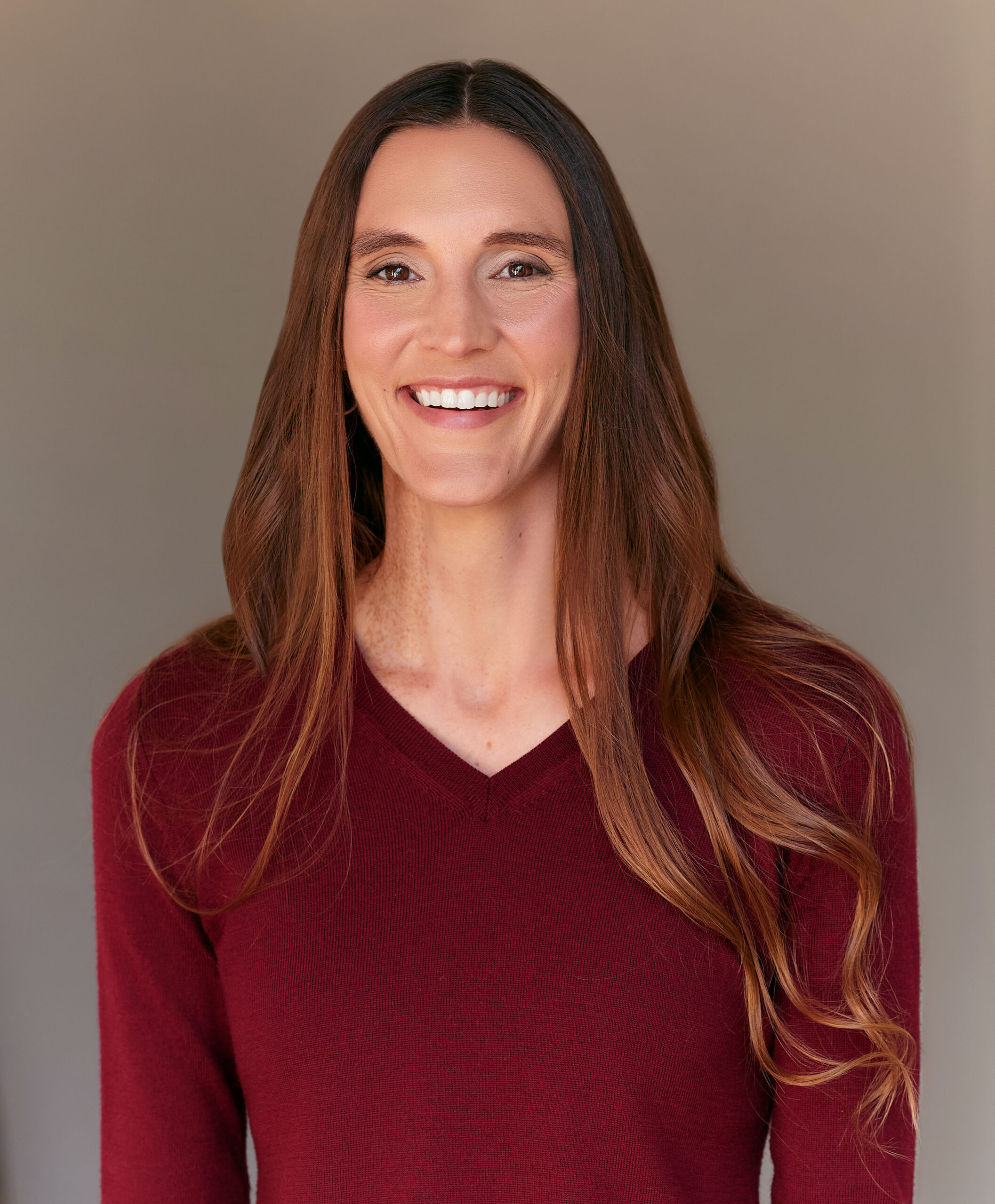 A smiling person with long, wavy brown hair is wearing a deep burgundy V-neck sweater. The background is a soft gray, complementing their warm expression.