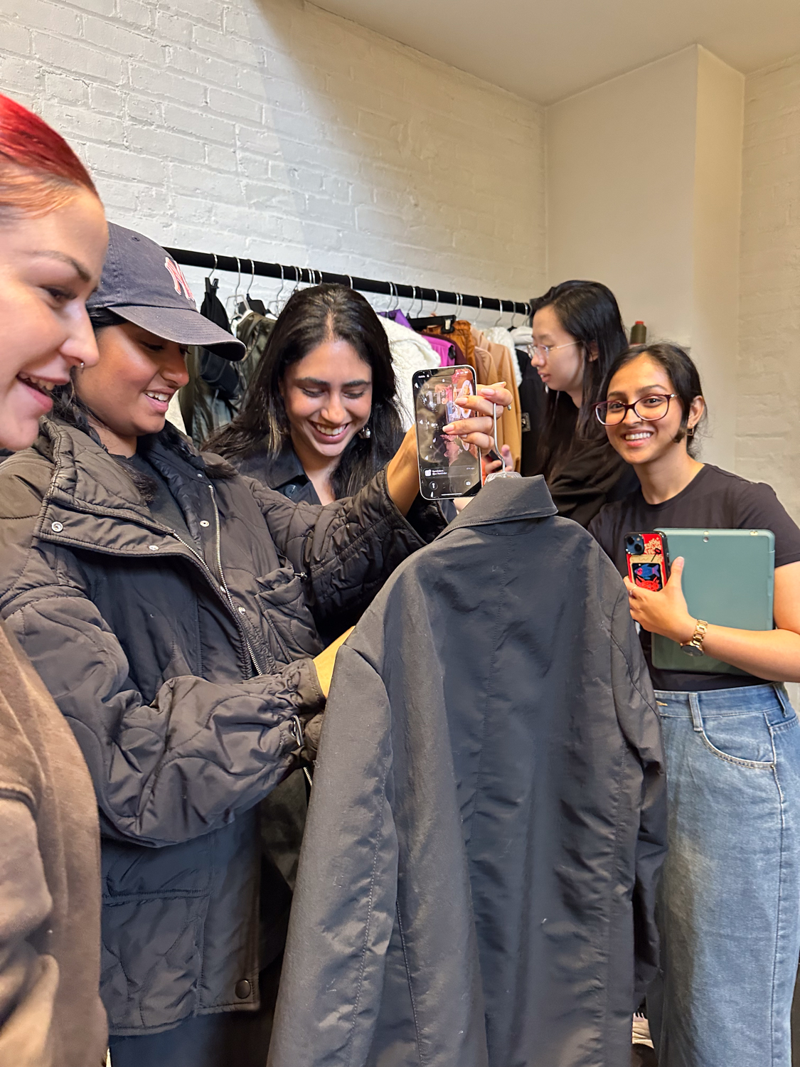 A group of five people are gathered in a clothing store, engaging with a dark jacket. Two people are smiling as they look at a phone, while another is holding the jacket. The setting features a clothing rack in the background with various garments hanging. The walls are white, creating a bright and casual atmosphere.