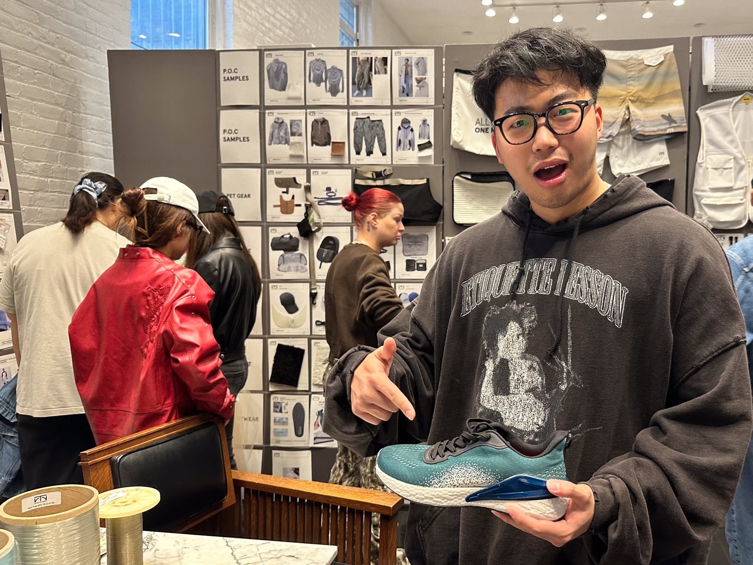 A young person wearing glasses and a dark hoodie stands in a store, pointing at a sneaker in their hand. The sneaker features a blue and white design. In the background, several people are browsing clothing items displayed on a wall. The setting has a modern, casual vibe with various apparel samples visible.