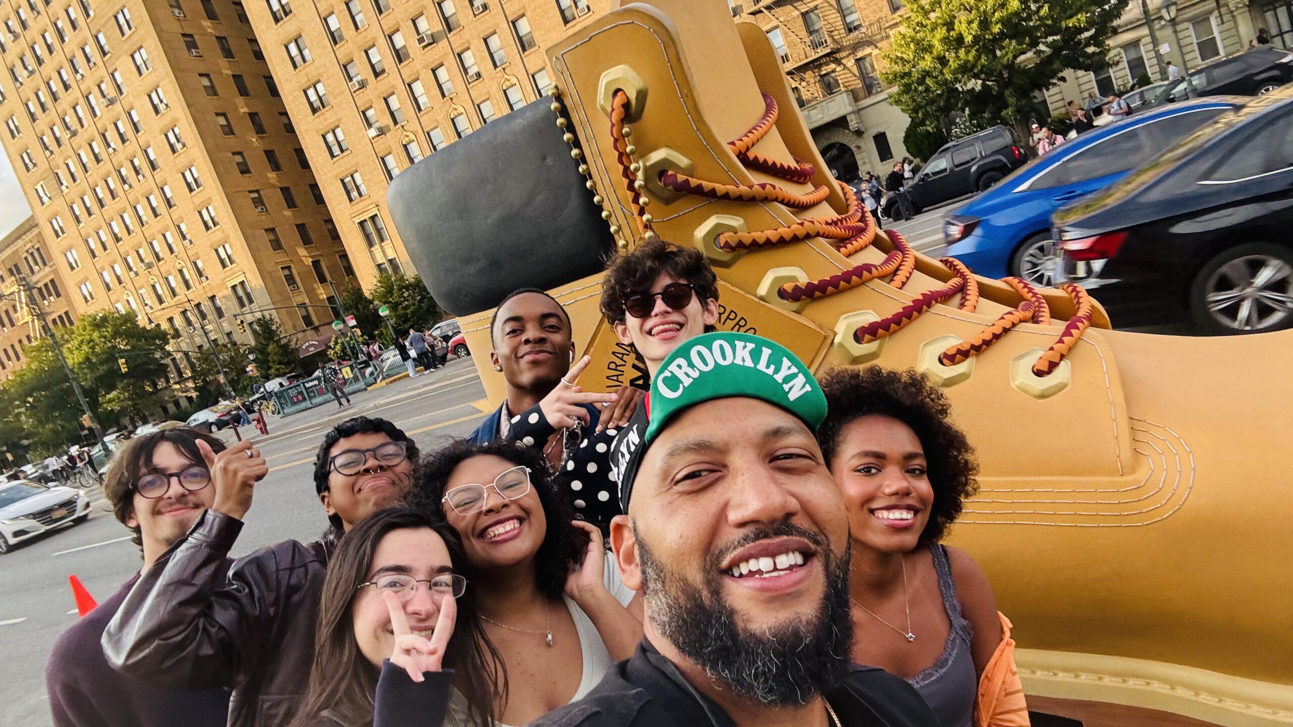 A group of seven young people poses for a selfie in front of a giant yellow boot statue. The setting is urban, with buildings and cars visible in the background. The individuals are smiling and making playful gestures, wearing casual clothing and accessories like hats and glasses. Trees and other pedestrians are also seen in the environment, contributing to a lively atmosphere.