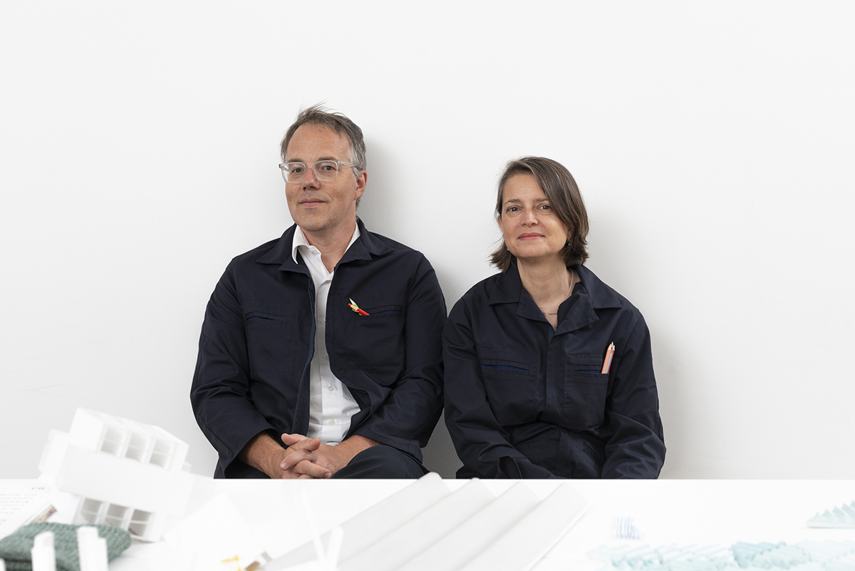 A man and a woman sit side by side against a plain white wall. Both are dressed in dark blue work jackets. The man has short gray hair and glasses, while the woman has shoulder-length brown hair. They appear relaxed and are seated in front of a table with various architectural models and materials.