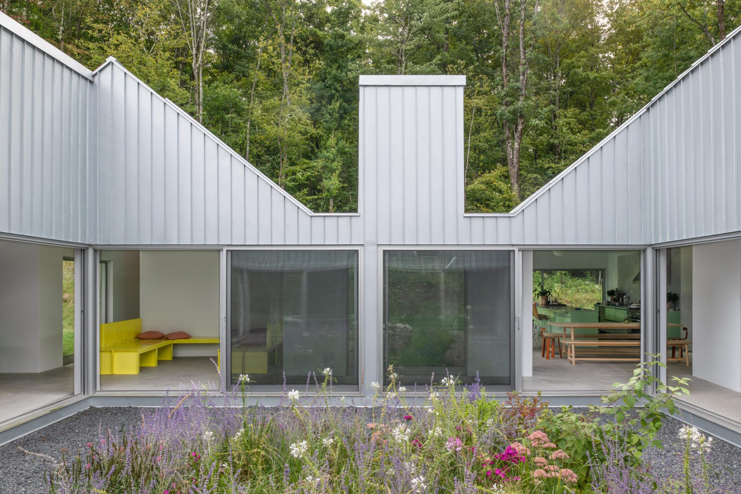 A modern house with a unique, sloped metallic roof design. The facade features large glass sliding doors that open to a spacious interior, showcasing a bright yellow seating area and a green kitchen area visible through the windows. A garden with colorful flowers, including lavender and pink blooms, is situated in the foreground, framed by gravel pathways and surrounding trees.