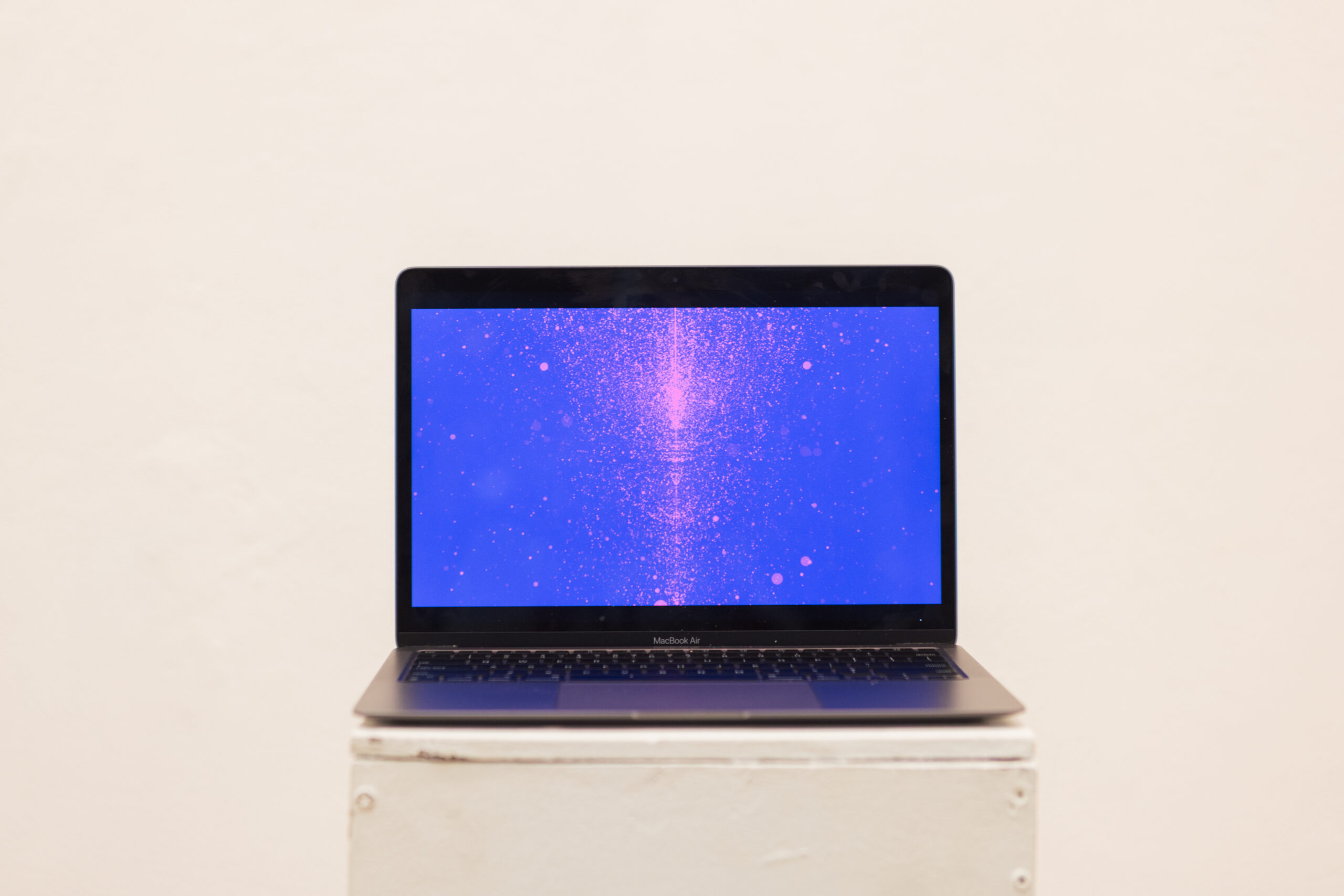 A MacBook Air placed on a white pedestal. The screen displays a vibrant blue background with pink particles or shapes scattered across it, creating an abstract visual effect. The wall behind the laptop is plain and light-colored.