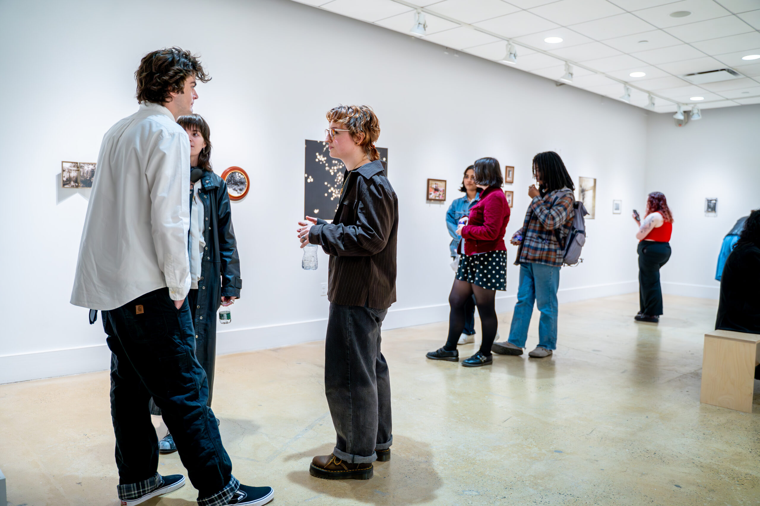 Visitors stand and converse inside a white-walled gallery during a photography exhibition. In the foreground, three people face one another in discussion, one holding a clear water bottle. Additional visitors are spaced along the gallery wall in the background, viewing small framed artworks and wall-mounted pieces. The room features polished concrete floors, overhead track lighting, and evenly spaced artworks arranged at eye level, creating an open, informal exhibition environment.