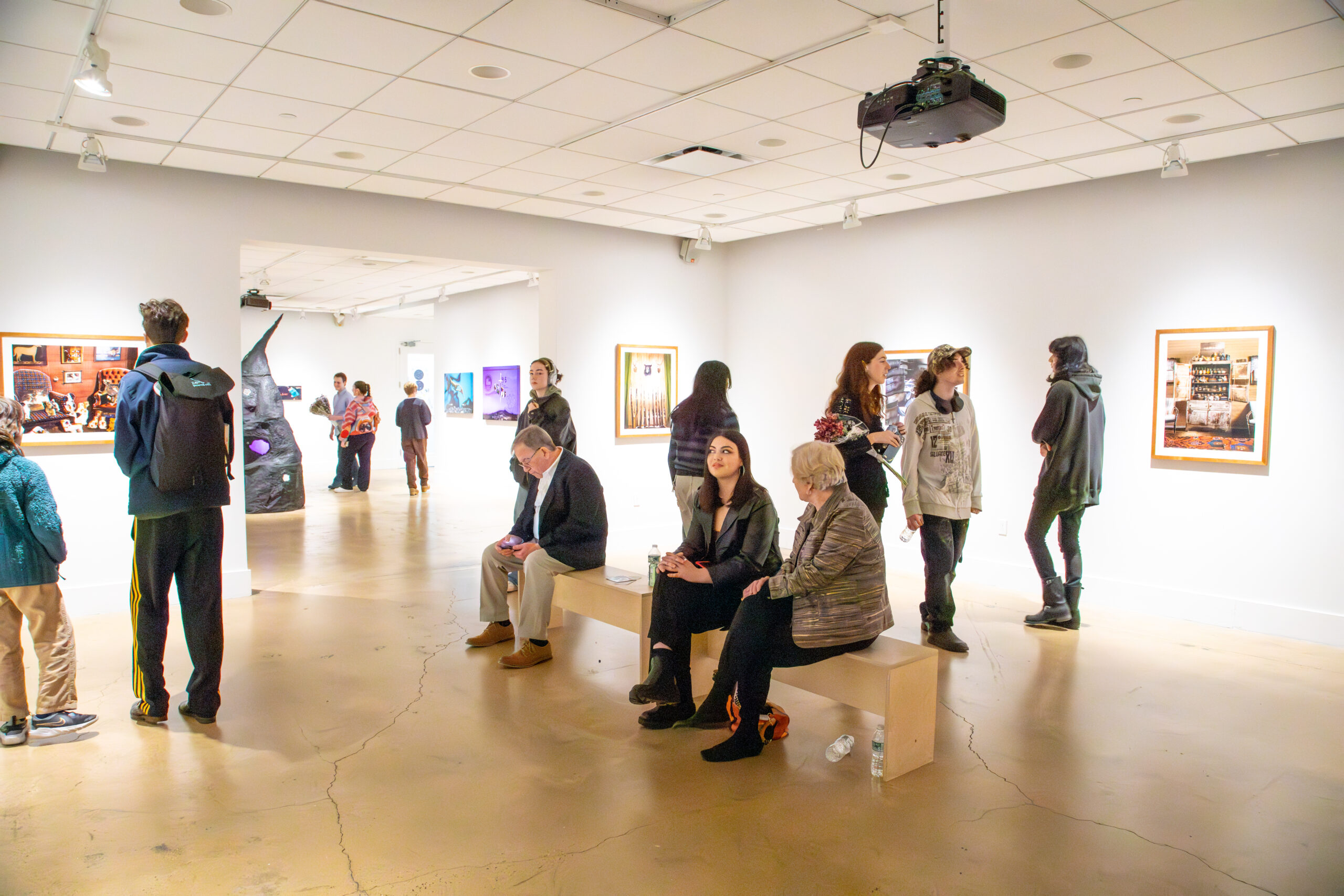 A wide view of a gallery exhibition space with multiple visitors dispersed throughout the room. Several people sit on a central wooden bench while others stand and converse or view framed photographic works mounted on white walls. A sculptural installation with an irregular vertical form is visible in the background near an open doorway connecting adjacent rooms. Overhead track lighting and a ceiling-mounted projector illuminate the space, emphasizing the active, social atmosphere of an art exhibition setting.