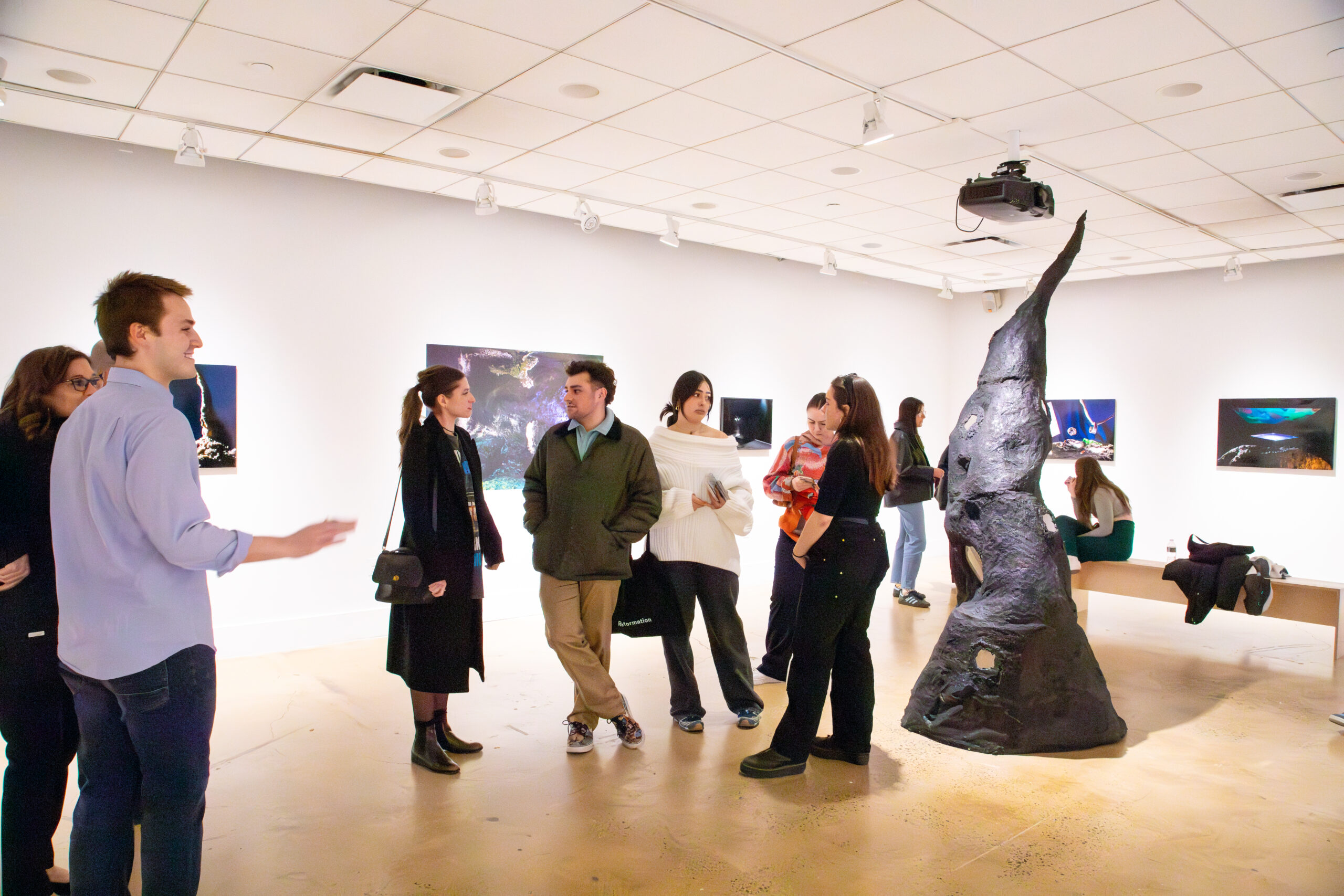 Visitors gather and converse inside a white-walled gallery during a photography exhibition. In the center of the room, a group of people stand in discussion near a tall, irregular dark sculptural form. Framed photographic works are mounted evenly along the walls behind them. Additional visitors are visible throughout the space, including some seated on a wooden bench. The gallery features polished concrete floors, overhead track lighting, and an open layout that supports movement, viewing, and informal conversation.