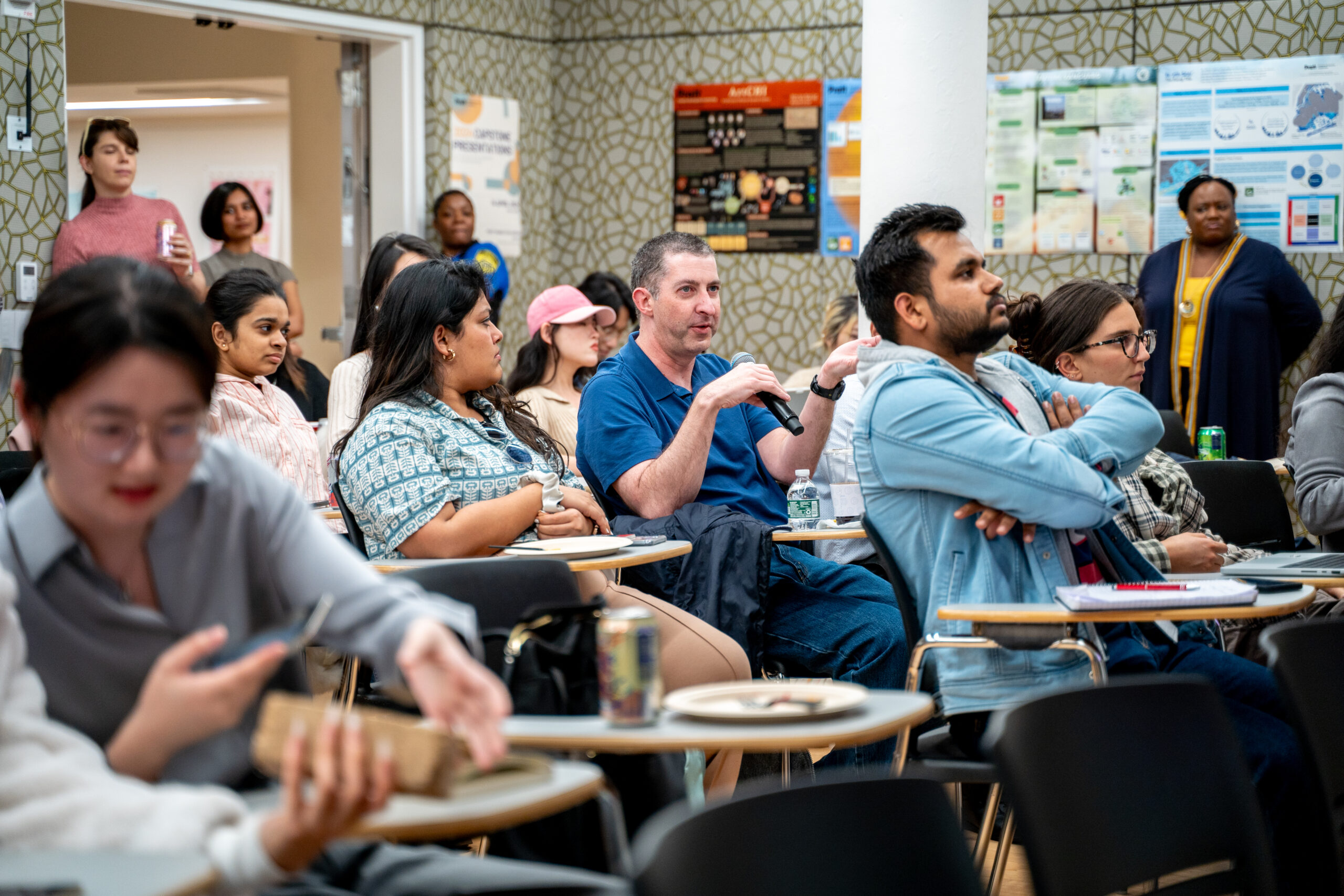 A classroom discussion with several adults seated at individual desks. In the center, a man wearing a blue polo shirt holds a microphone and speaks while gesturing with his hand. Around him, students of diverse backgrounds listen attentively, some with notebooks, drinks, or laptops on their desks. Posters and informational displays are visible on the patterned wall behind them, and additional attendees stand near the doorway observing the session.