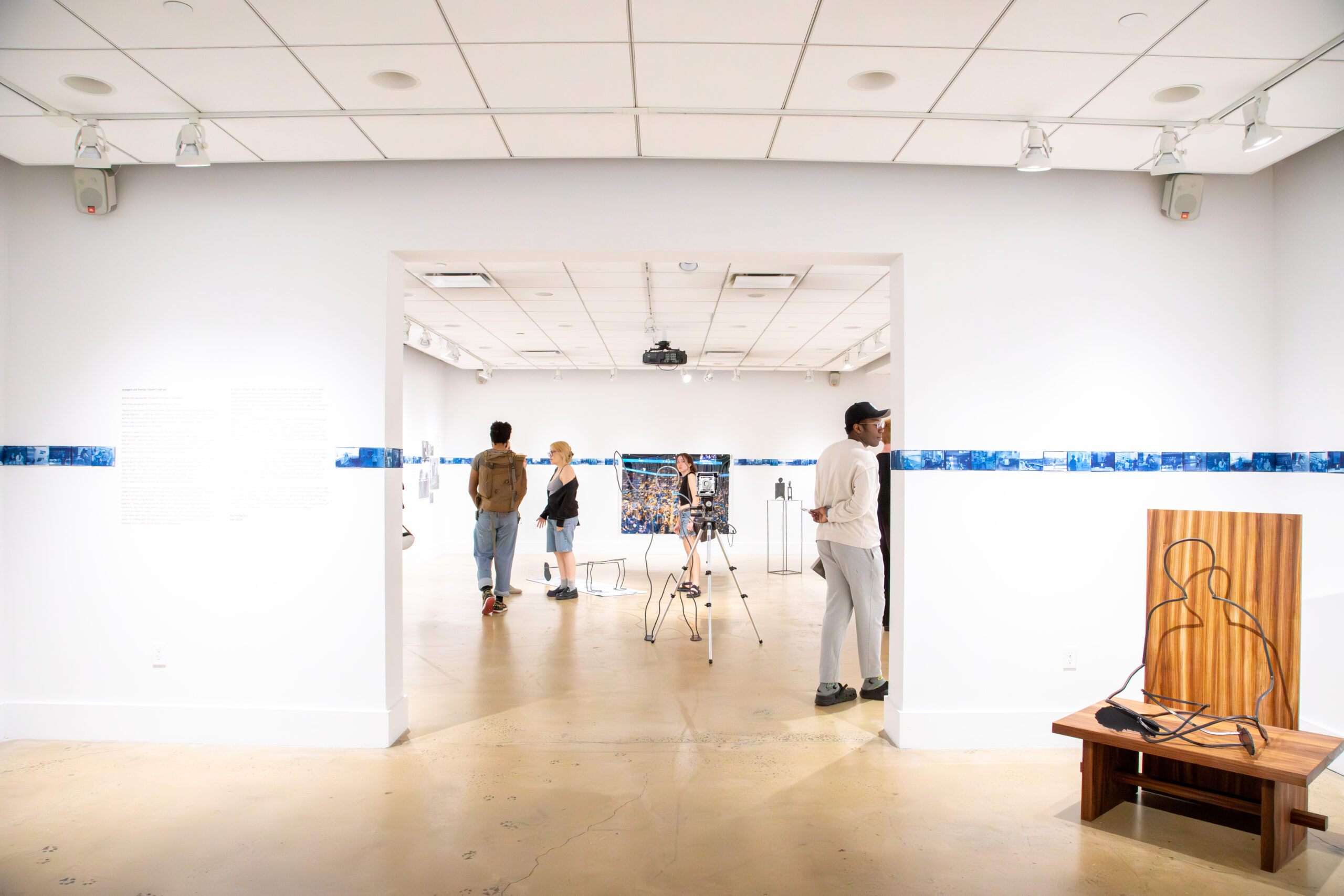Wide view of a contemporary art gallery with white walls and polished concrete floors. Several visitors stand and walk through the space, viewing artworks installed along the walls and in the central room beyond a large rectangular doorway. A horizontal band of small blue photographic prints runs along the walls at eye level. In the foreground at right, a wooden bench supports a sculptural metal outline of a seated human figure. A camera on a tripod is set up in the central gallery, suggesting documentation or exhibition recording. Overhead track lighting and ceiling panels illuminate the space evenly.
