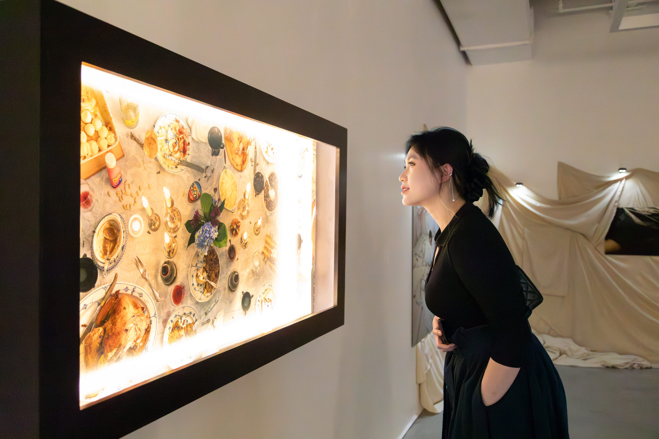 A gallery visitor stands in profile, leaning slightly forward to view an illuminated wall-mounted lightbox artwork. The artwork displays a tabletop scene with plates of food, candles, glassware, and floral elements arranged in a ceremonial or communal setting, glowing from internal lighting and framed in black. The visitor wears a dark long-sleeved dress with pockets and has their hair pulled back, one hand resting near their midsection. In the background, a softly lit installation of draped fabric occupies the gallery space. The environment features white walls, a polished floor, and overhead gallery lighting.