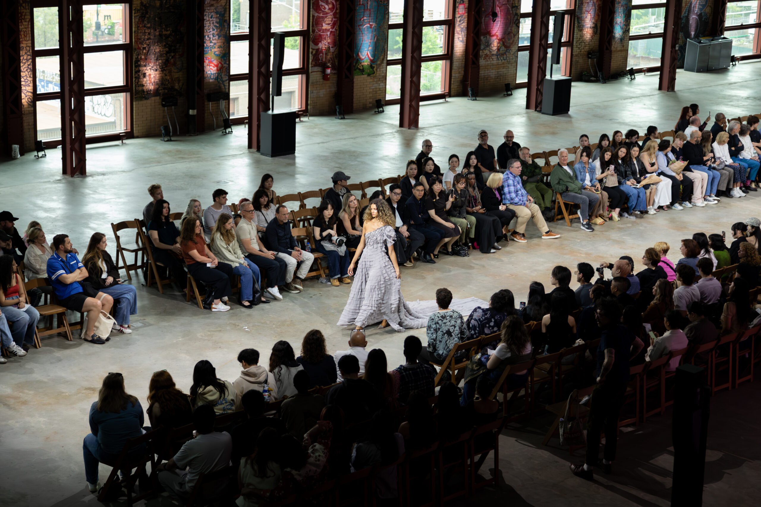 Wide view of an indoor fashion runway show in a large industrial hall, with audience members seated in rows on both sides as a model in a tiered gray gown walks down the runway.