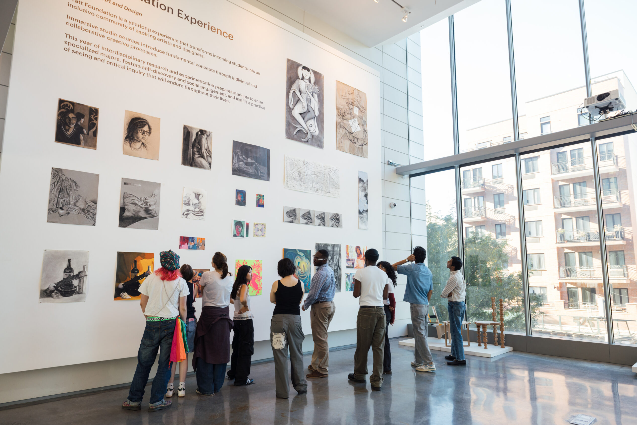 A diverse group of people stands in front of a large wall displaying various artworks, including sketches and paintings. They appear engaged and are examining the pieces. The space is bright with large windows allowing natural light in, and a portion of an urban landscape is visible outside. The wall features a heading about an art program, and artworks include figures, still lifes, and abstract designs.
