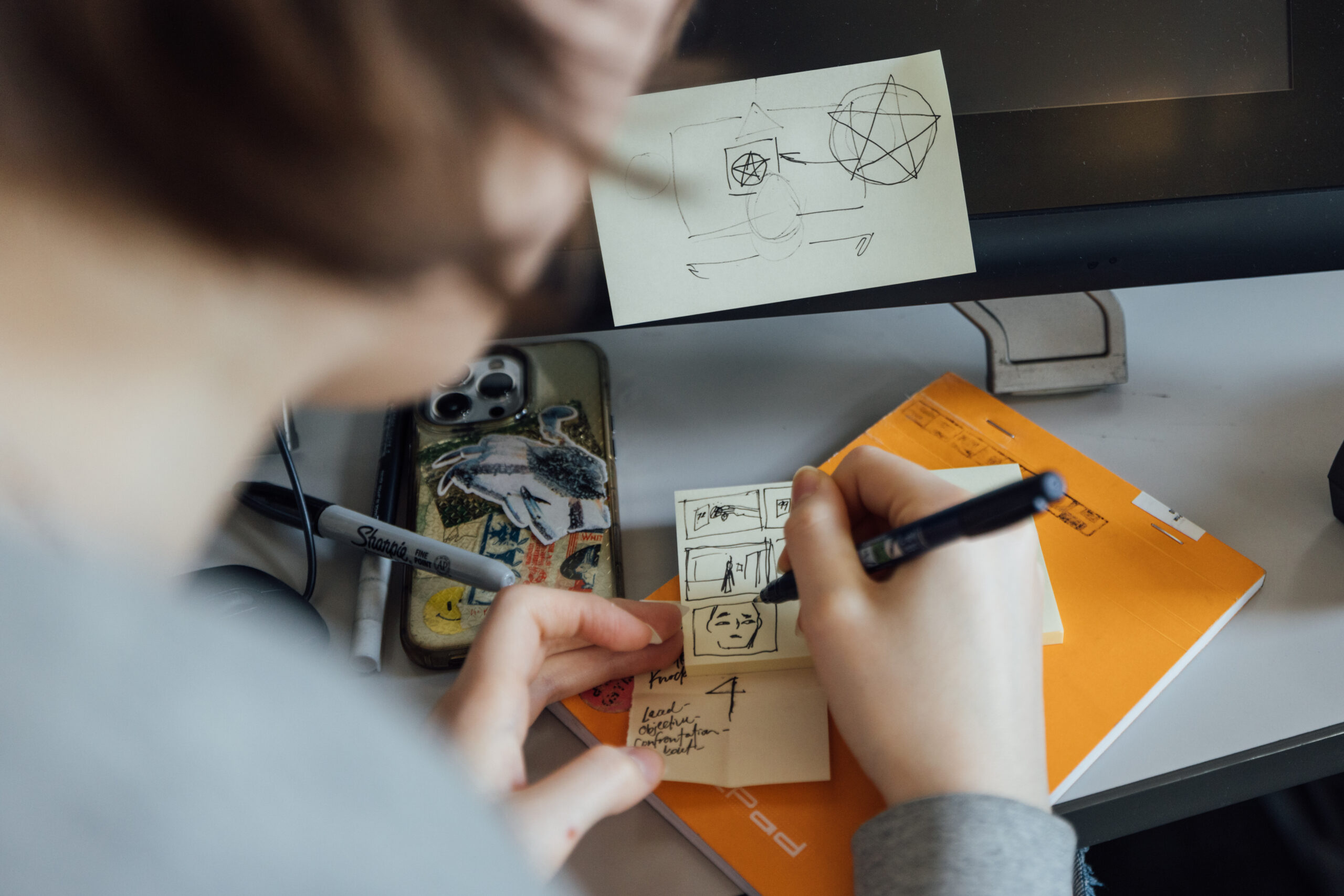 Close-up of a person sketching small storyboard frames on a sticky note with a black pen at a desk. The drawings show simple scene panels and a face. On the desk are an orange notebook, additional sticky notes with handwritten notes, a smartphone with stickers on the case, and a Sharpie marker. A computer monitor in the background has another sticky note attached with rough diagram sketches.