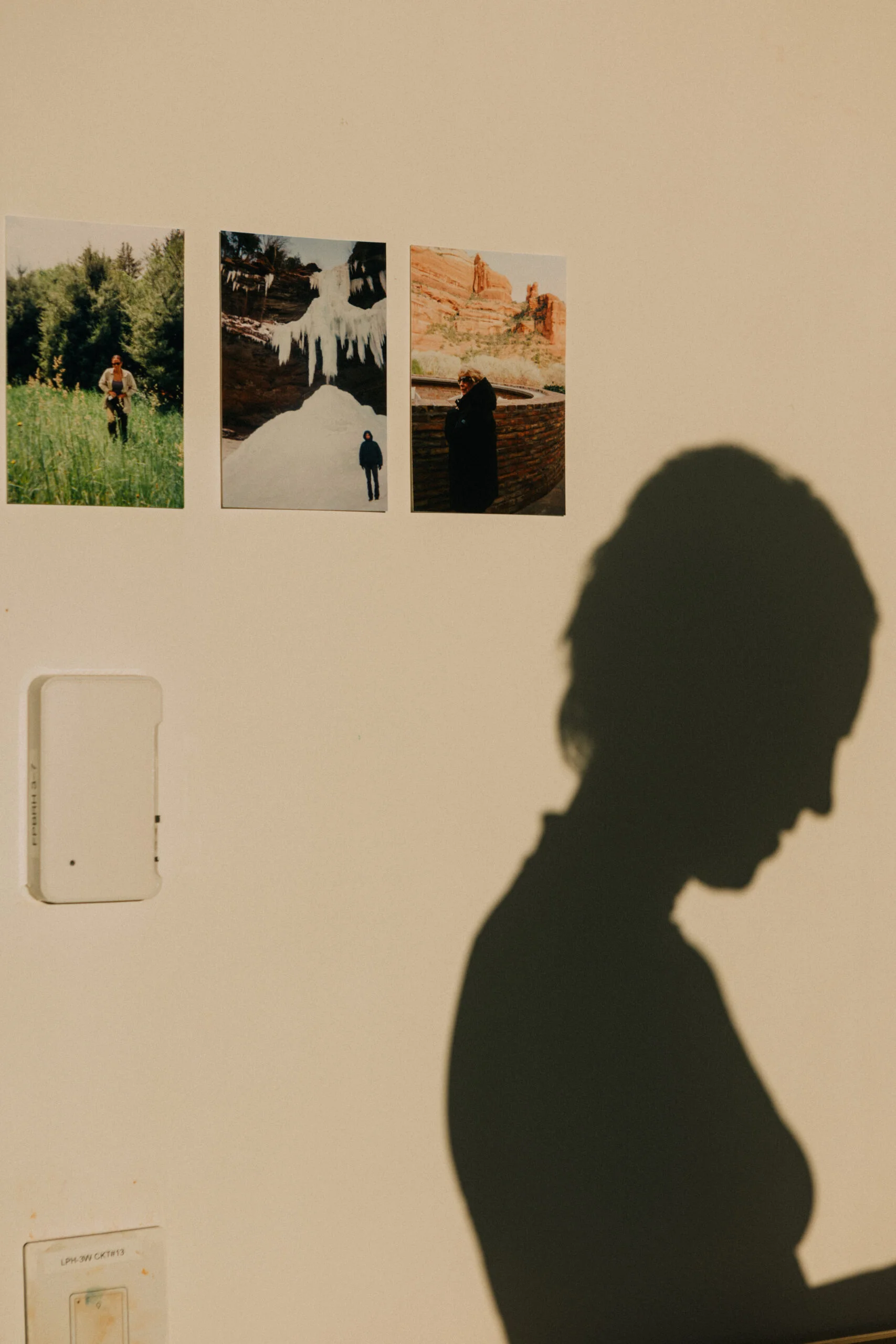 A wall with three photos hanging: one shows a person in a green field, the second features icicles and a person in a dark coat, and the third captures a person standing near a stone wall with red rock formations in the background. A shadow of a person is visible on the wall, facing downward, with their hair pulled back. A light switch and a small white object are also present near the bottom left.