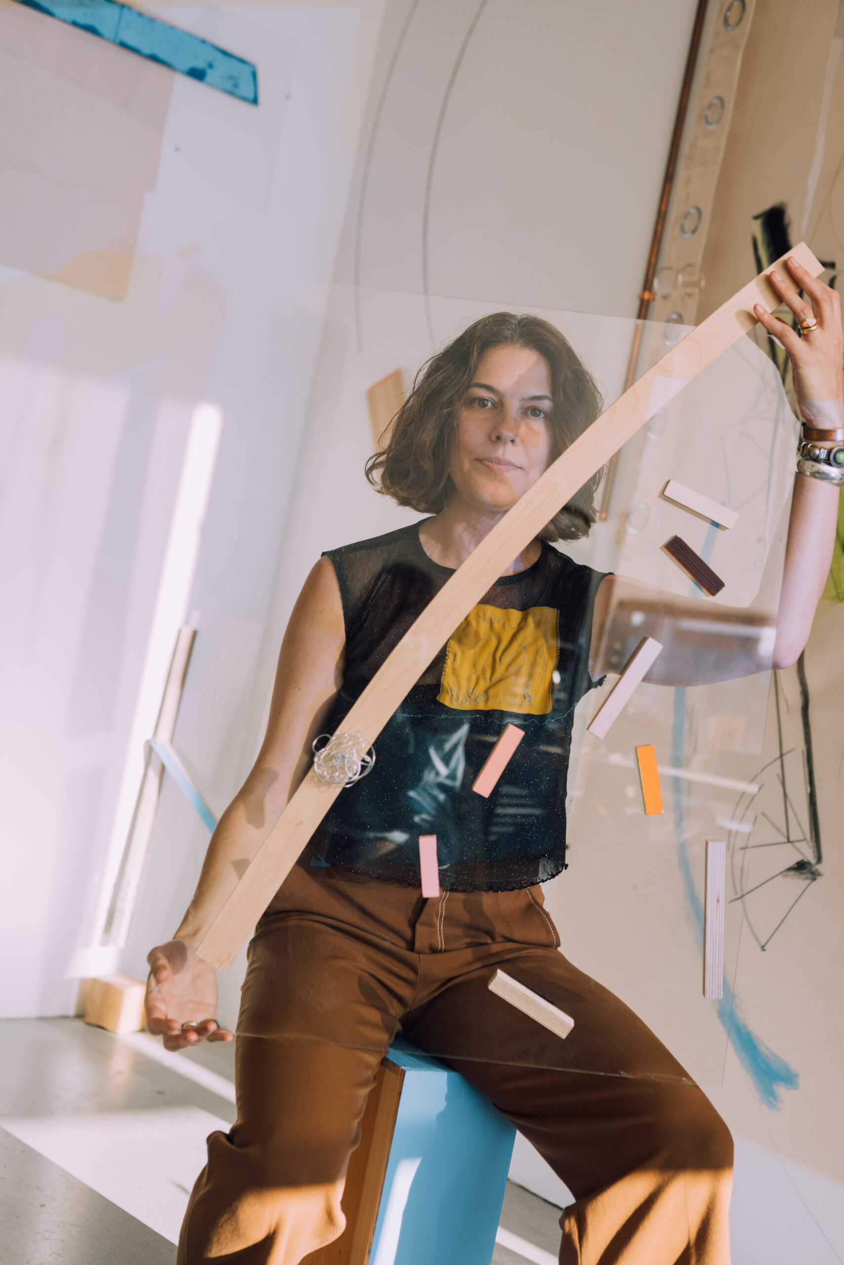 A woman sits on a light blue block, holding an arch-shaped transparent object with various colored wooden pieces attached to it. She wears a black top with a yellow square and brown pants. The background features abstract art and soft lighting, creating a creative studio atmosphere.