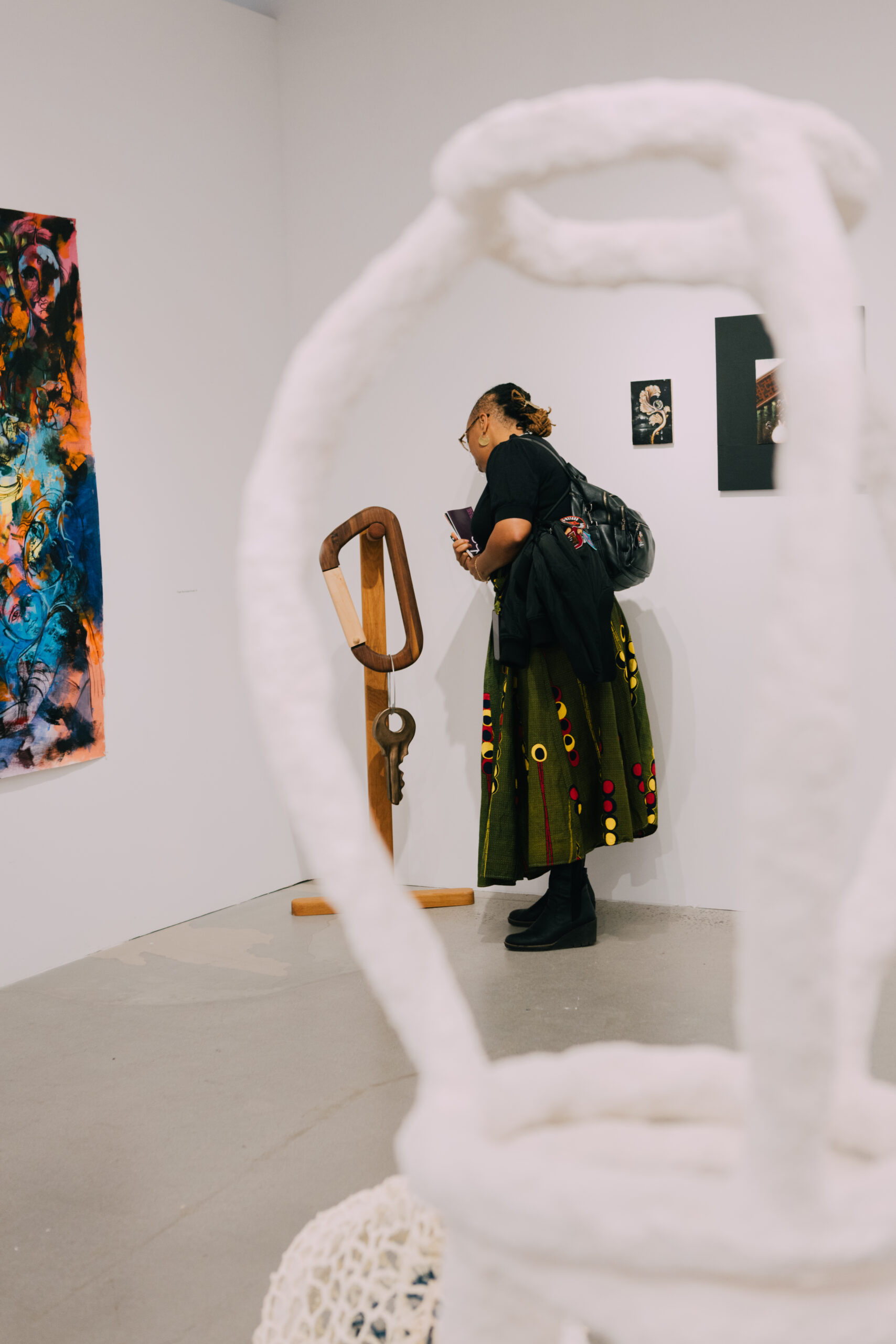 A person with a dark shirt and a multi-colored skirt stands next to a wooden sculpture in an art gallery. They hold a notebook and are facing the artwork. In the foreground, a white, sculptural form partially frames the scene, and colorful abstract paintings are visible on the wall behind them.