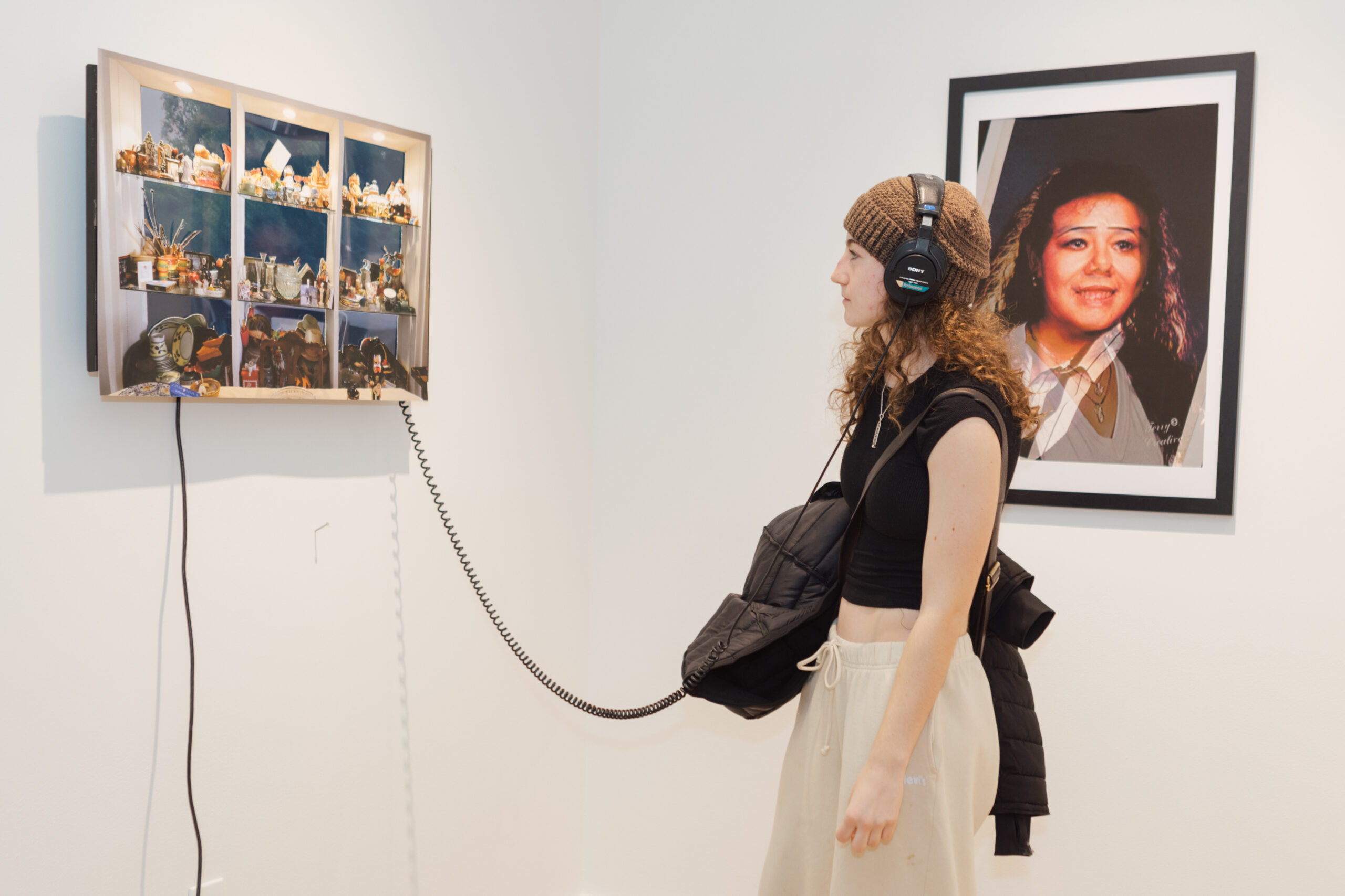 A gallery visitor stands in profile wearing over-ear headphones connected by a cord to an illuminated wall-mounted artwork. The person wears a knit beanie, sleeveless black top, light-colored pants, and carries a jacket over one arm. The artwork consists of a framed lightbox displaying multiple small objects arranged in compartments, suggesting a shrine or collection. To the right, a framed photographic portrait of a woman hangs on the wall. The gallery space has white walls, minimal fixtures, and even overhead lighting.