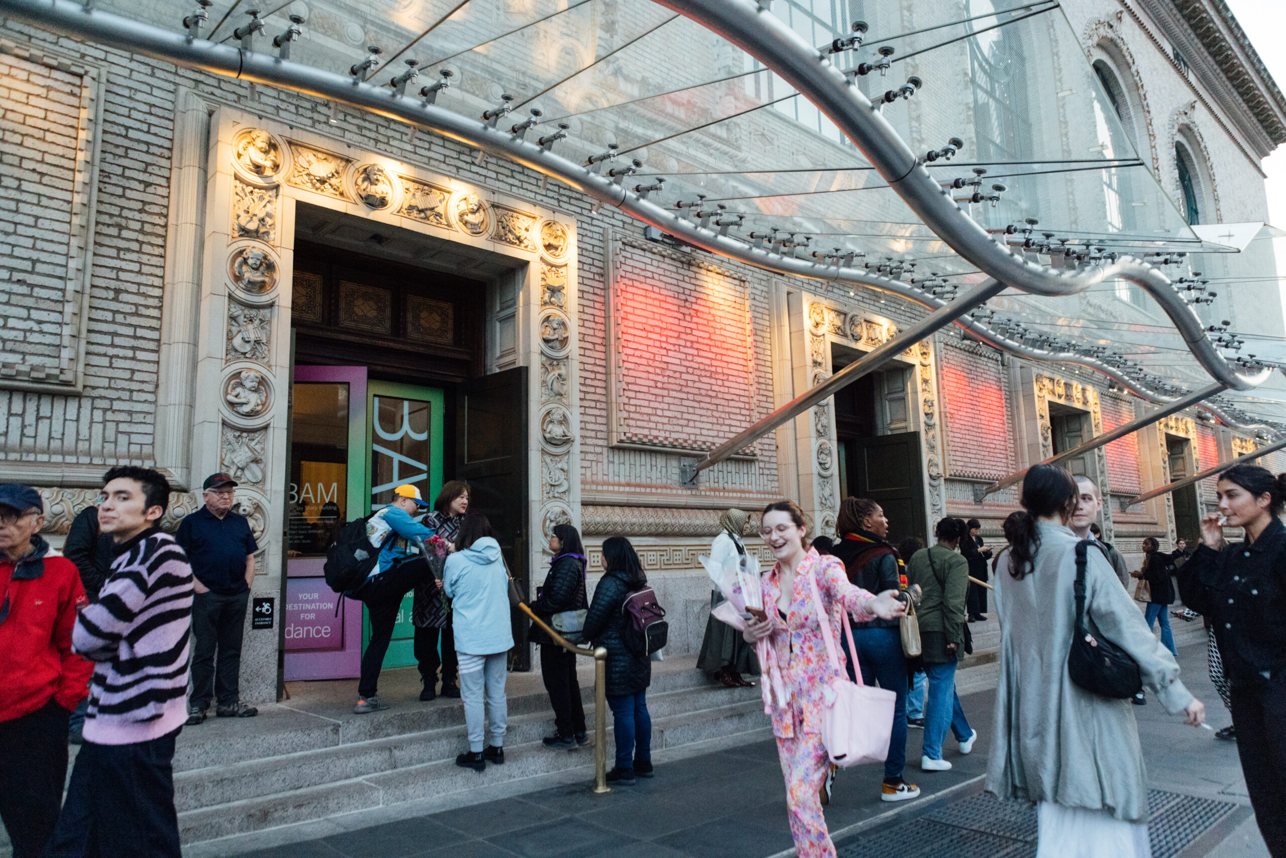 Crowd gathered outside a building entrance beneath a curved glass-and-metal canopy, with people walking and talking on the sidewalk near open doors and decorative stonework.