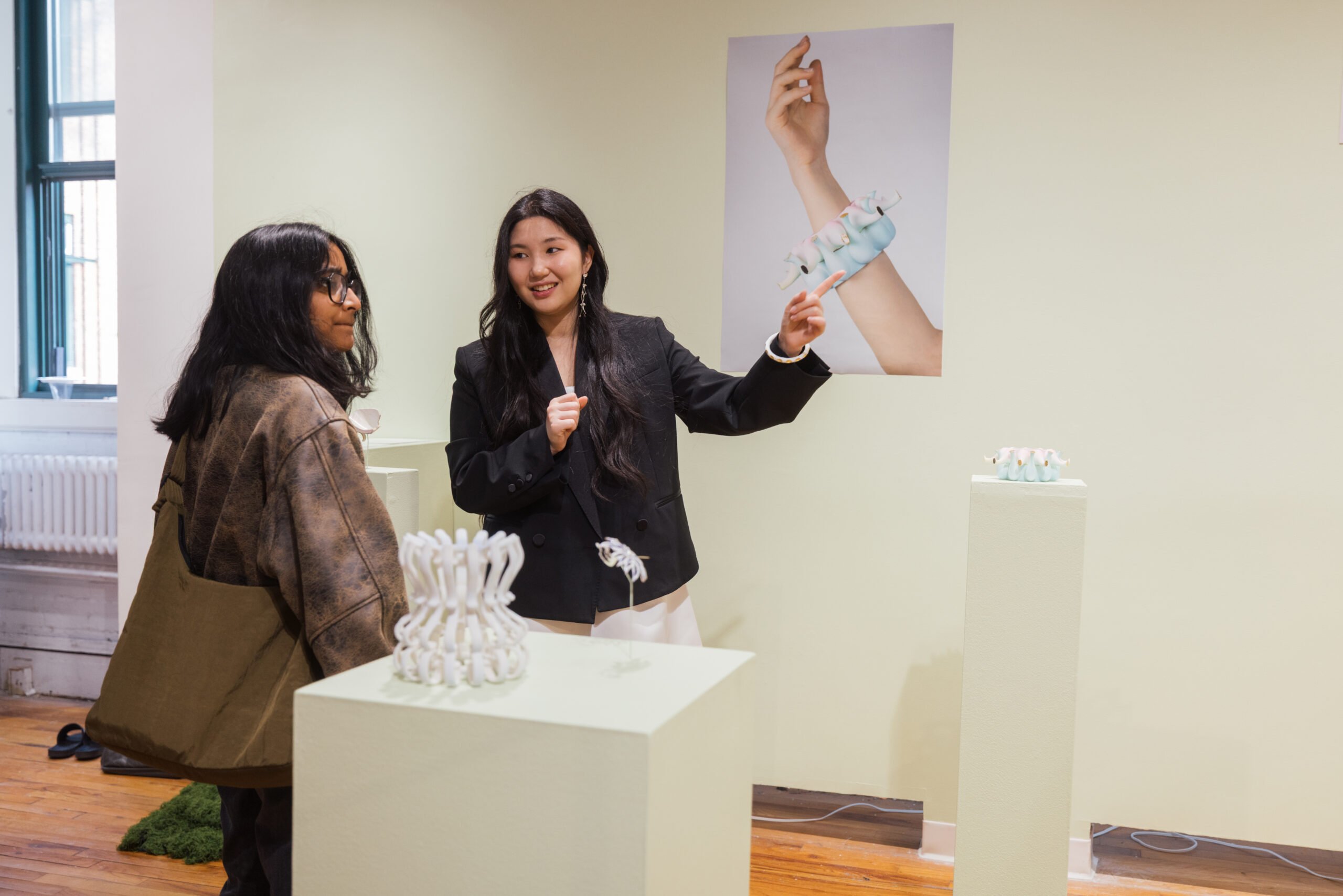 Two people talk in a gallery exhibition space beside display pedestals with small sculptural objects, while one person gestures toward a wall poster showing an arm wearing a jewelry-like piece.