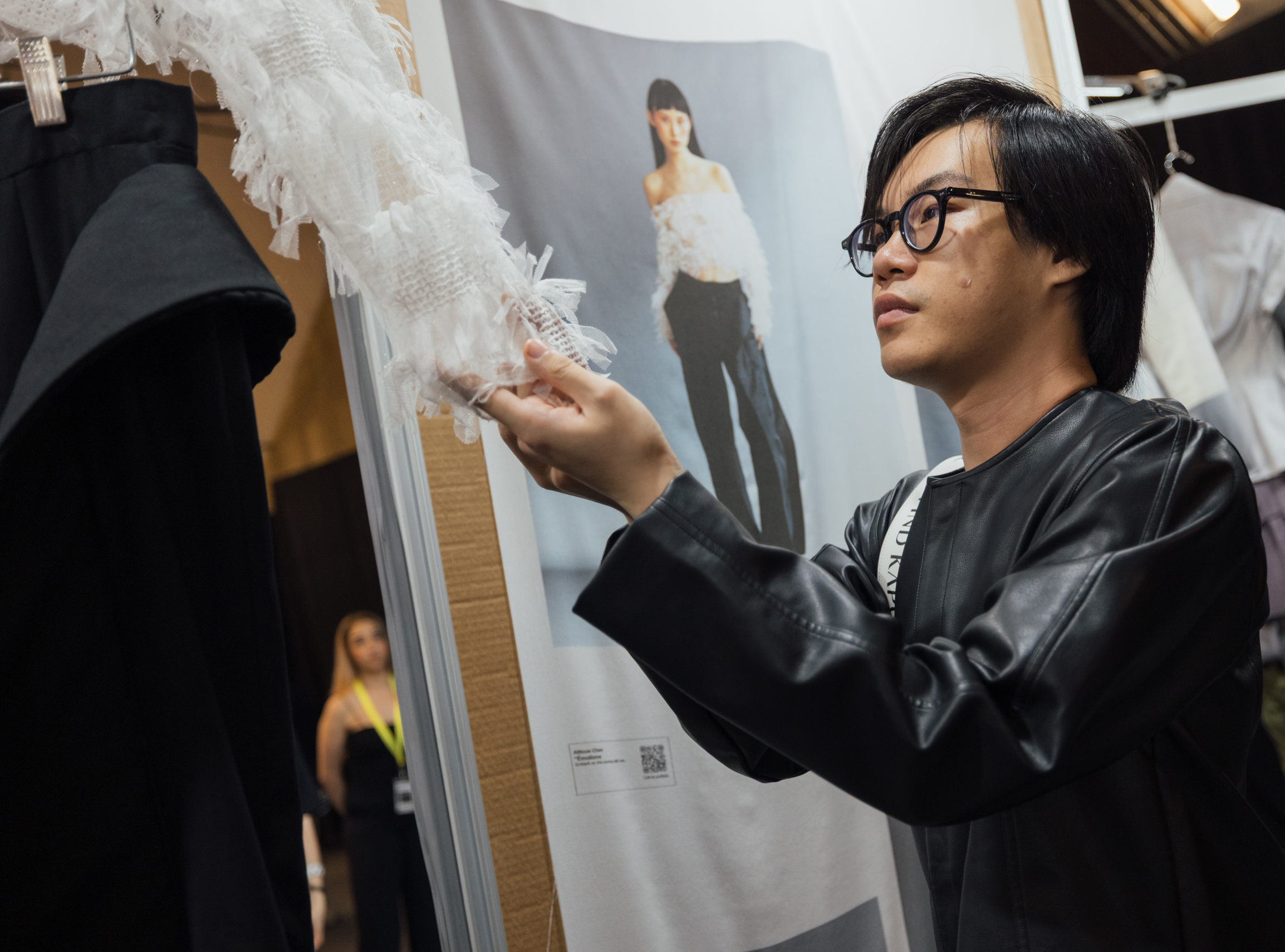 A young man wearing a black leather jacket examines a clothing piece with ruffled white sleeves in a fashion exhibit. Behind him, a poster displays a model wearing an off-the-shoulder top and dark pants. Another person can be partially seen in the background. The setting appears to be a fashion show or showcase event.