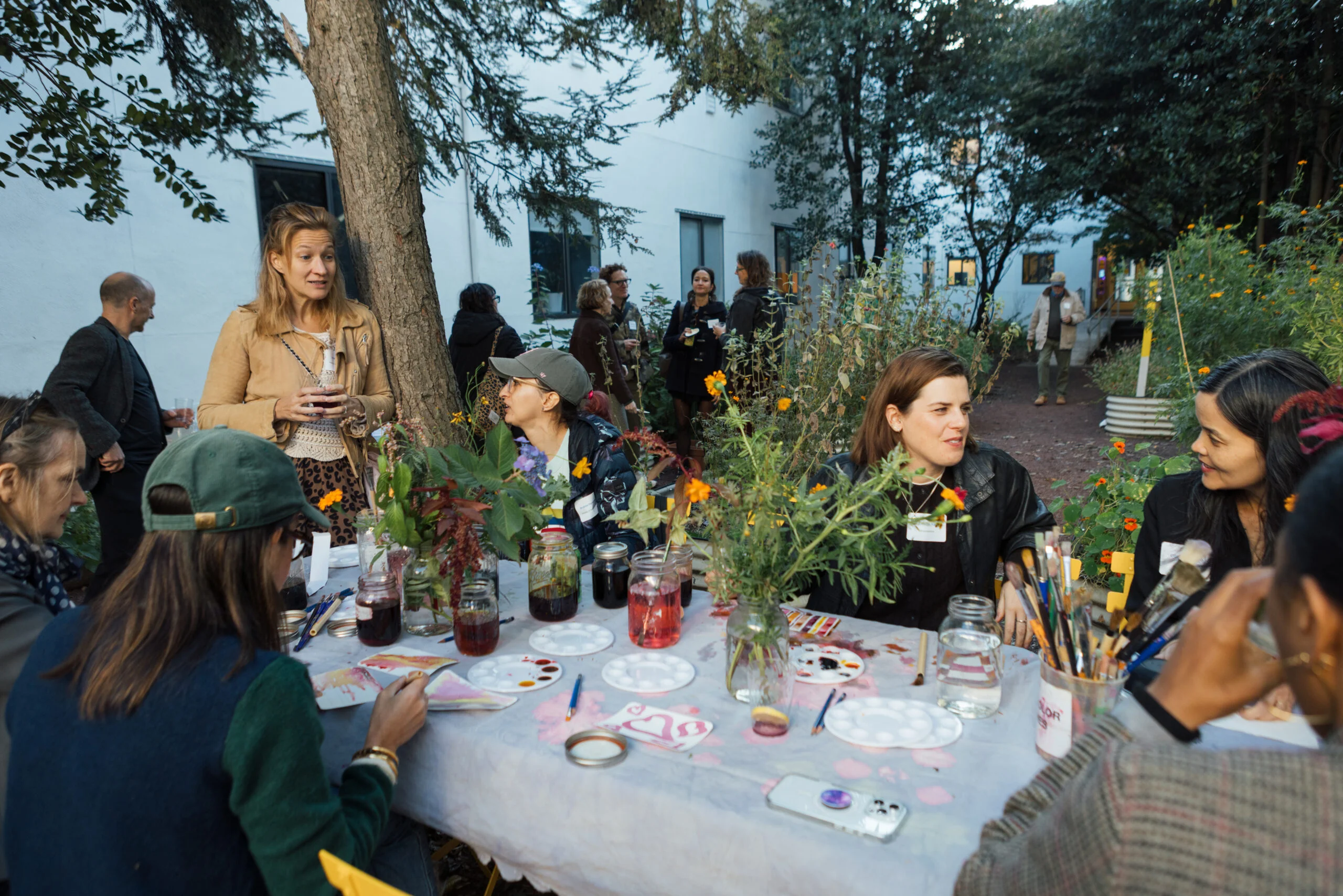 A group of people are gathered around a table in a garden setting, engaged in a creative activity. Some individuals are painting while others are discussing. The table is adorned with jars of colorful liquids, paint palettes, and various art supplies. In the background, additional people are mingling among trees and flowers, creating a lively atmosphere. The setting appears relaxed and collaborative, suggesting a communal artistic experience.