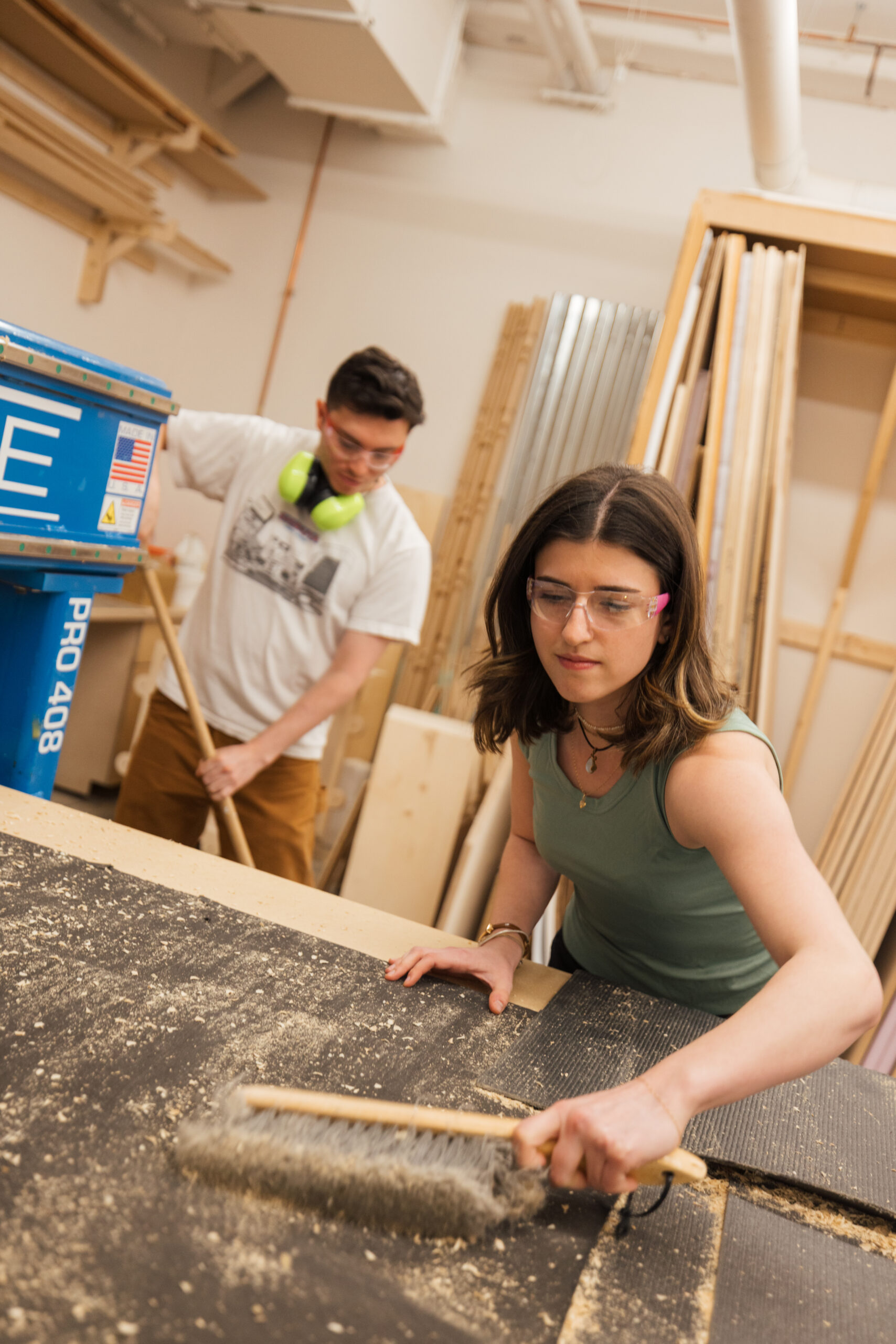 A young woman in a green tank top uses a broom to sweep up sawdust on a workbench. She wears glasses and a necklace. In the background, a young man wearing a white t-shirt and headphones leans over, holding a long handle as he works on the floor. The workshop is filled with wood materials and tools, indicating construction or carpentry activities.