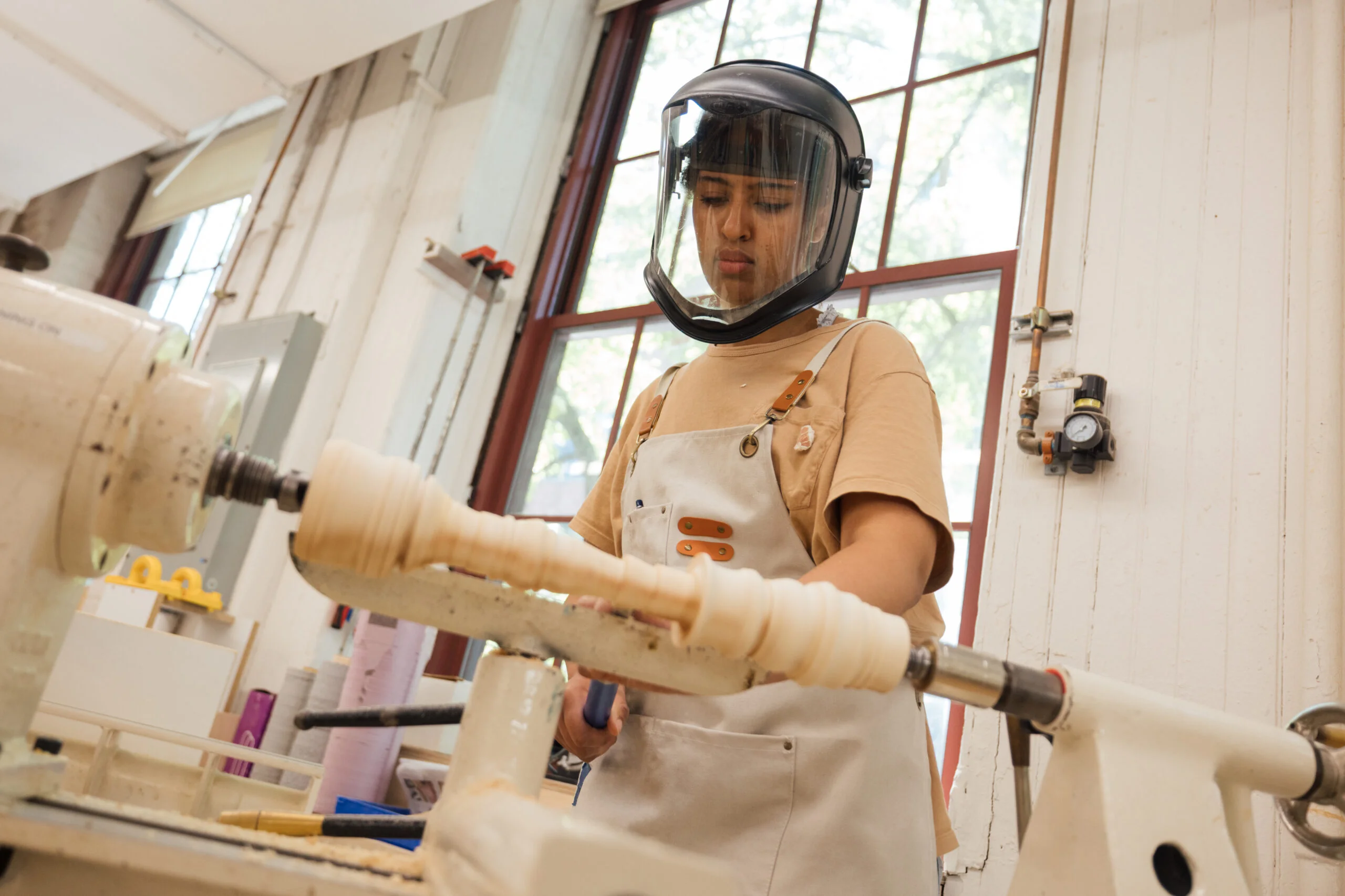 A person wearing a protective face shield and a beige apron is concentrating on operating a lathe in a bright workshop. The lathe is shaping a wooden piece with spiral grooves, and natural light streams through large windows in the background. Various tools and materials are visible on the workbench, contributing to the creative atmosphere of the space.