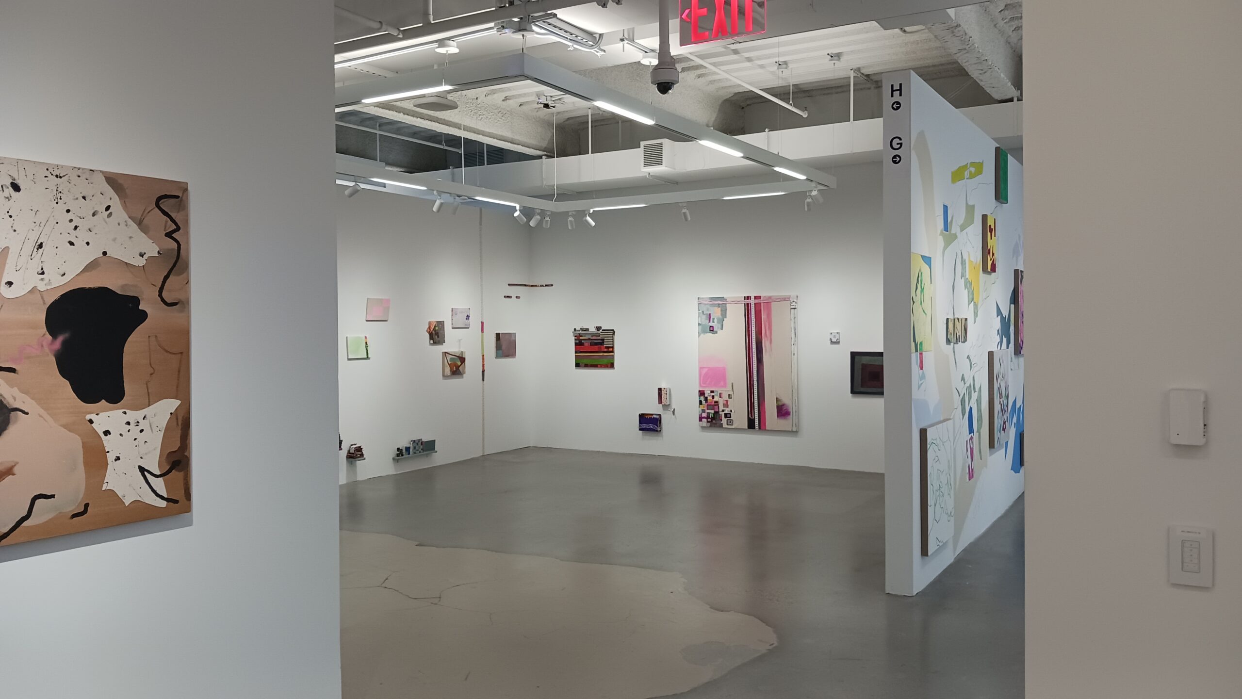 Wide view of a contemporary art gallery interior with white walls, polished concrete floors, and overhead track lighting. Multiple abstract and mixed-media artworks are installed on the walls, including paintings and small sculptural pieces. A partial wall frames the view into the main gallery space, and an illuminated red EXIT sign hangs from the ceiling, indicating an accessible public exhibition environment.
