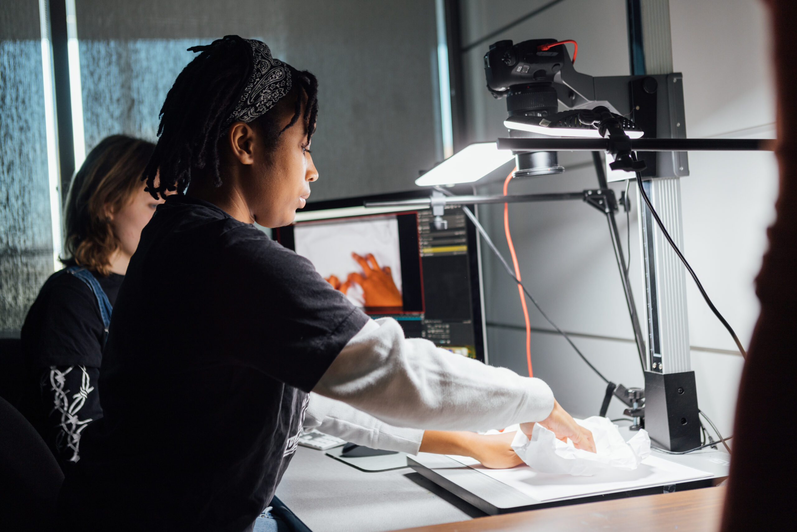 Two students work at a desk in a studio environment using a camera and lighting setup mounted above a flat work surface. The student in the foreground adjusts a crumpled white paper object beneath a mounted camera and bright light, suggesting stop-motion or animation production. A computer monitor behind them displays an image of an orange object in progress. The space includes studio equipment, cables, and controlled lighting, indicating a digital media or animation lab setting.