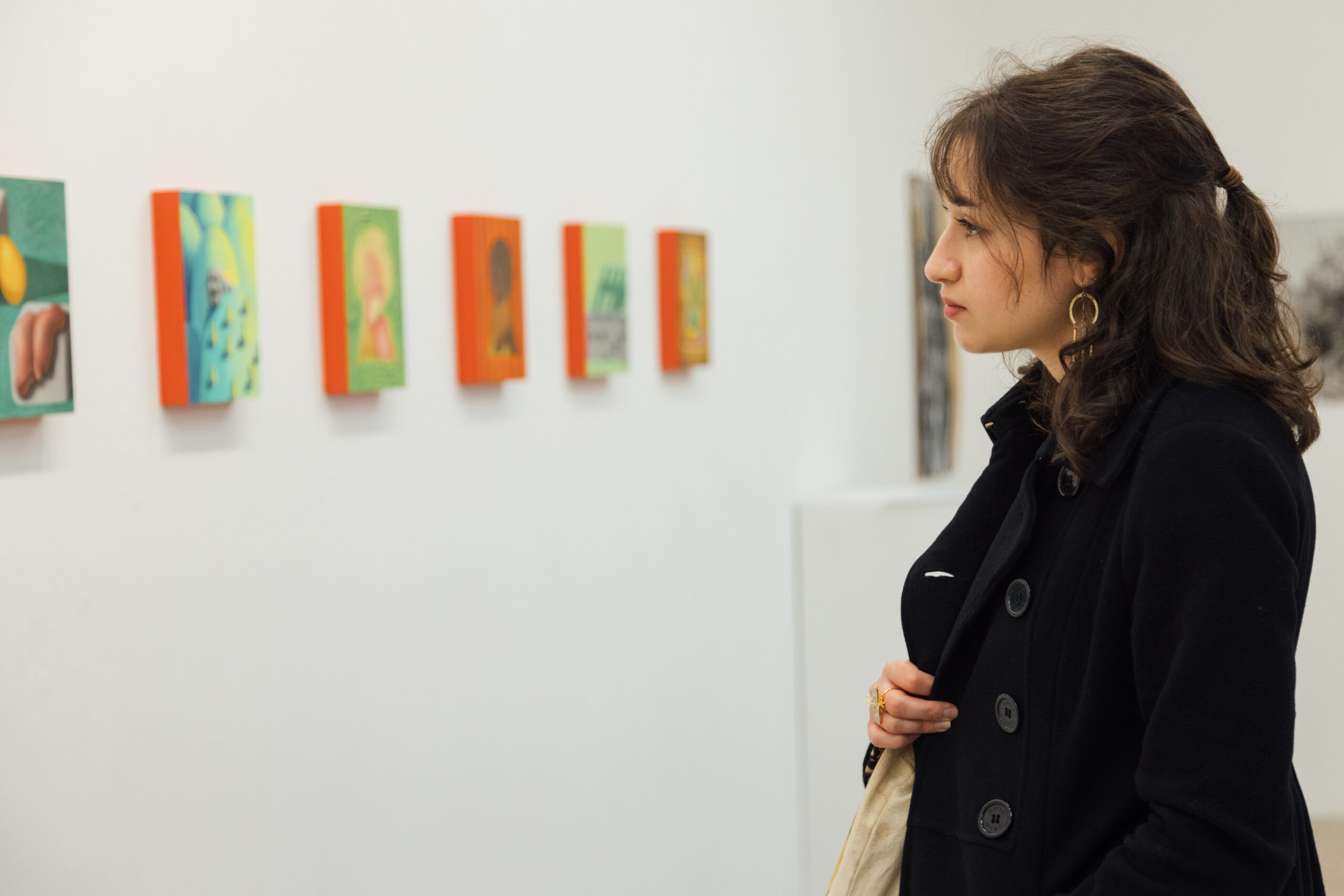 A visitor stands in profile inside a white-walled gallery, closely observing a row of small, colorful paintings mounted in a horizontal line on the wall. The person wears a dark coat and gold hoop earrings and holds a light-colored tote bag, appearing focused and contemplative. The artworks feature bright backgrounds with figurative and abstract elements, evenly spaced along the wall, suggesting a fine arts thesis exhibition setting.