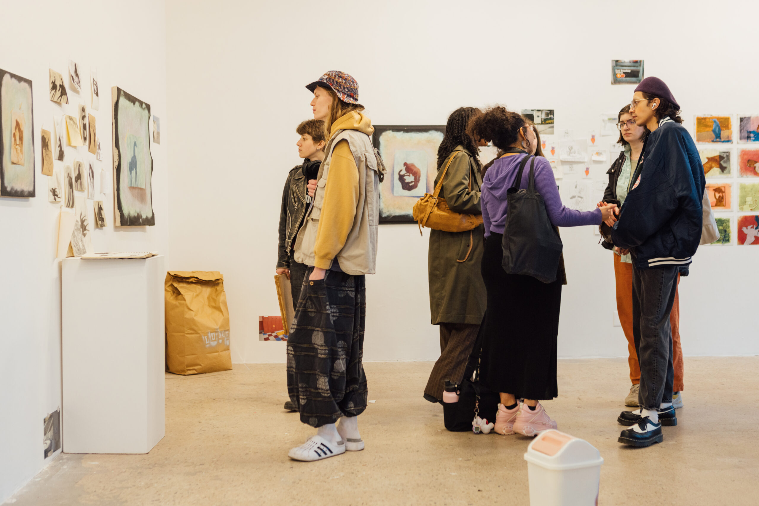 A group of visitors stands inside a white-walled gallery space during an art exhibition, viewing and discussing works displayed on the walls. On the left, small drawings and mixed-media pieces are arranged in a clustered installation above a low pedestal. Several people stand closely together in the center and right of the image, engaged in conversation while facing the artworks. The scene includes varied clothing styles, tote bags, and backpacks, with additional framed artworks visible in the background, indicating an active thesis exhibition environment.