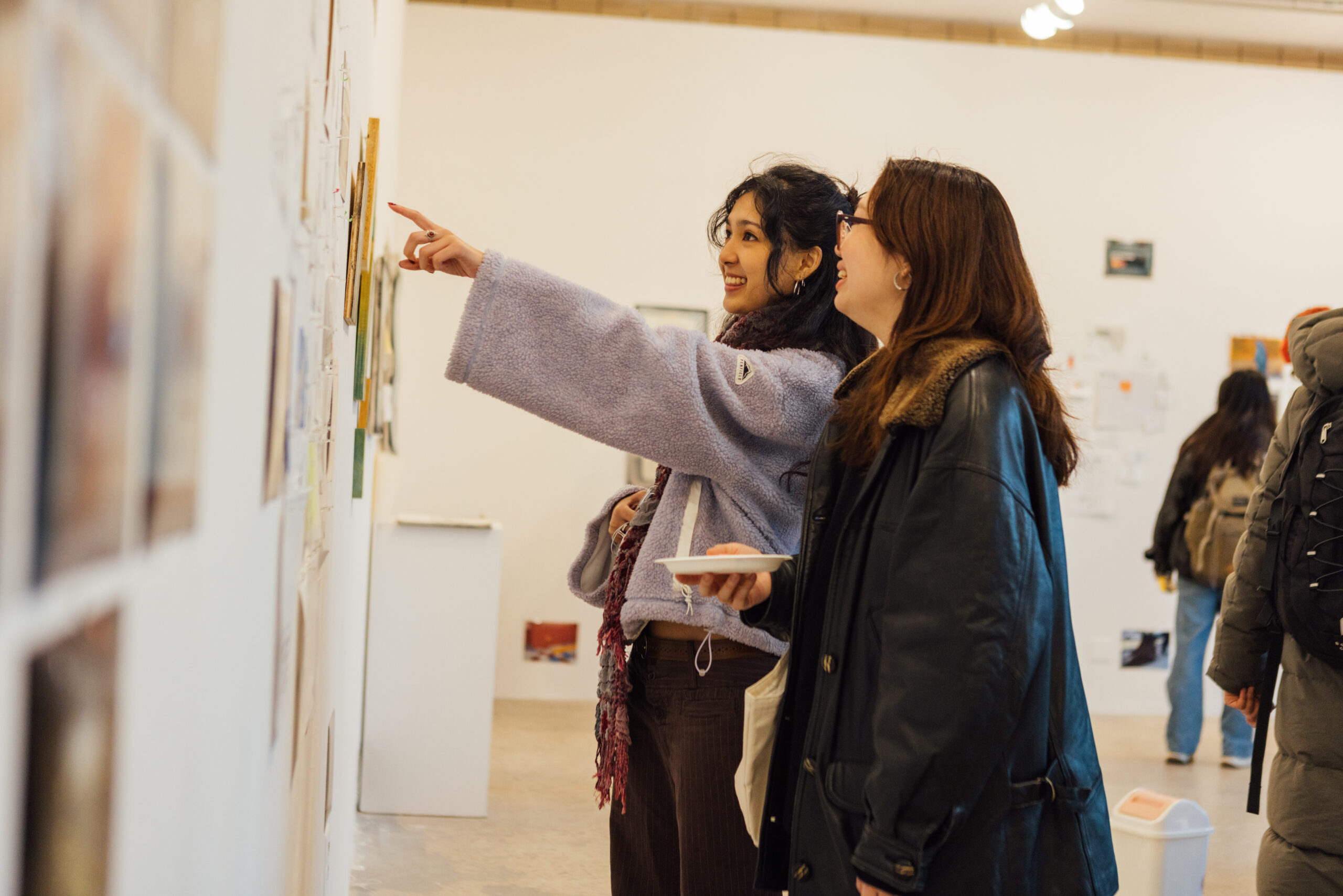 Two visitors stand side by side in a white-walled gallery, smiling and engaging with a wall-mounted display of drawings and mixed-media works. One person gestures with an outstretched arm, pointing toward a specific artwork, while the other looks on attentively, holding a small plate. Both wear winter jackets, and other visitors and artworks are visible in the background, indicating an active BFA drawing thesis exhibition setting.