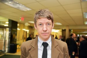 Head-and-shoulders portrait of a person standing indoors in a lobby or reception area. The person faces the camera with a neutral expression and wears a tan blazer, light-colored dress shirt, and black tie. The background is softly blurred, showing overhead fluorescent ceiling panels, warm interior lighting, and several people conversing in business attire.