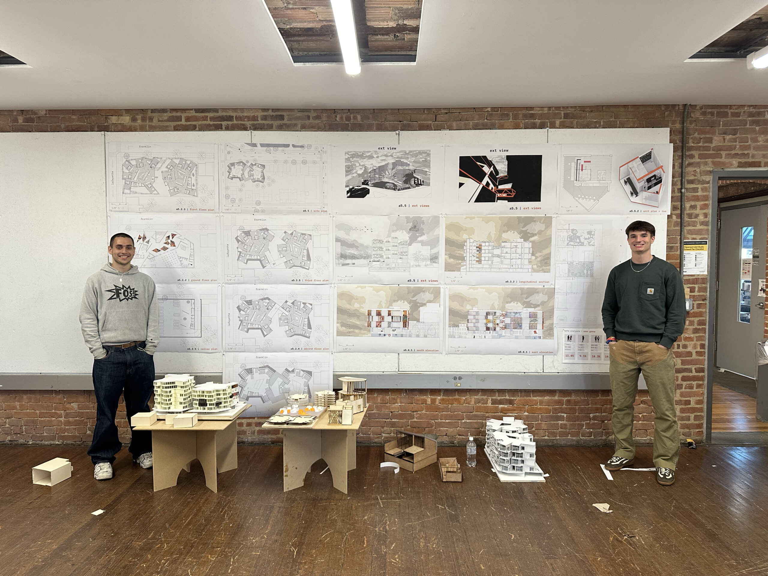 Two individuals stand in a room with a brick wall, surrounded by architectural drawings pinned on a board. They are next to wooden tables displaying architectural models. One person wears a gray hoodie and jeans, while the other wears a dark green sweatshirt and khaki pants. Various scale models and a water bottle are visible on the tables and floor.
