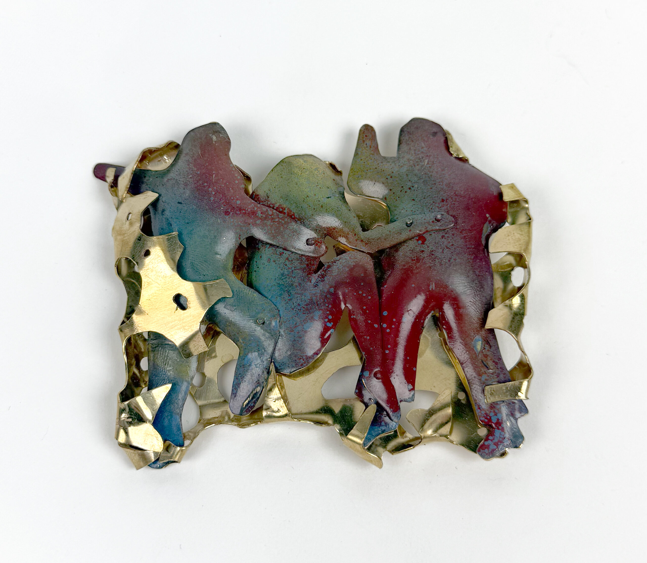 A colorful brooch featuring abstract human figures intertwined, made of layered metal and painted in shades of red, blue, and gold. The figures are set against a textured, gold-toned background with cut-out shapes, enhancing the piece's depth and visual interest.