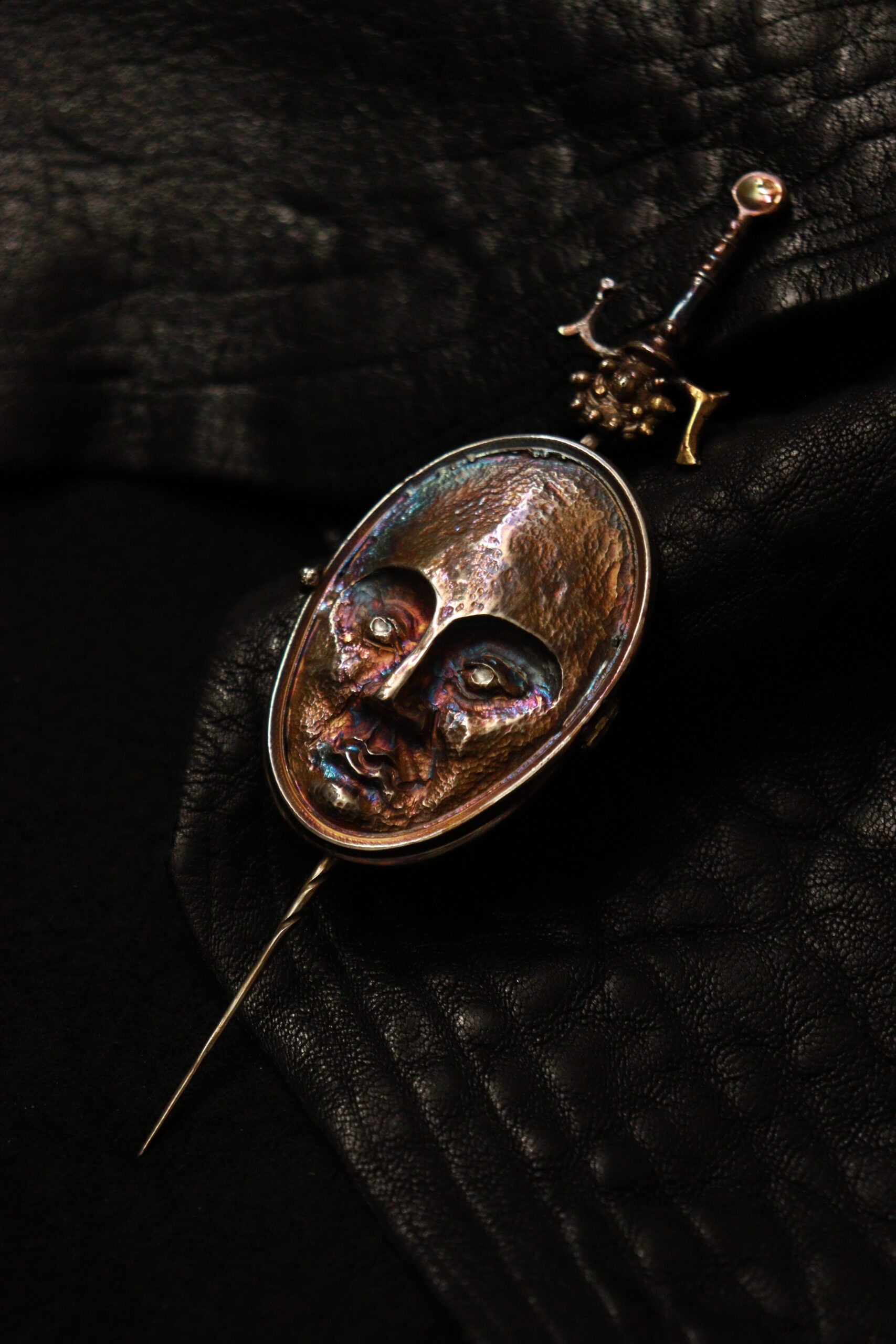A close-up image of a decorative brooch featuring an oval face with intricate, colorful detailing. The face has deep-set eyes and a textured surface, reflecting various hues. The brooch includes a thin pin and a small sword-like element at the top, set against a textured black leather background.