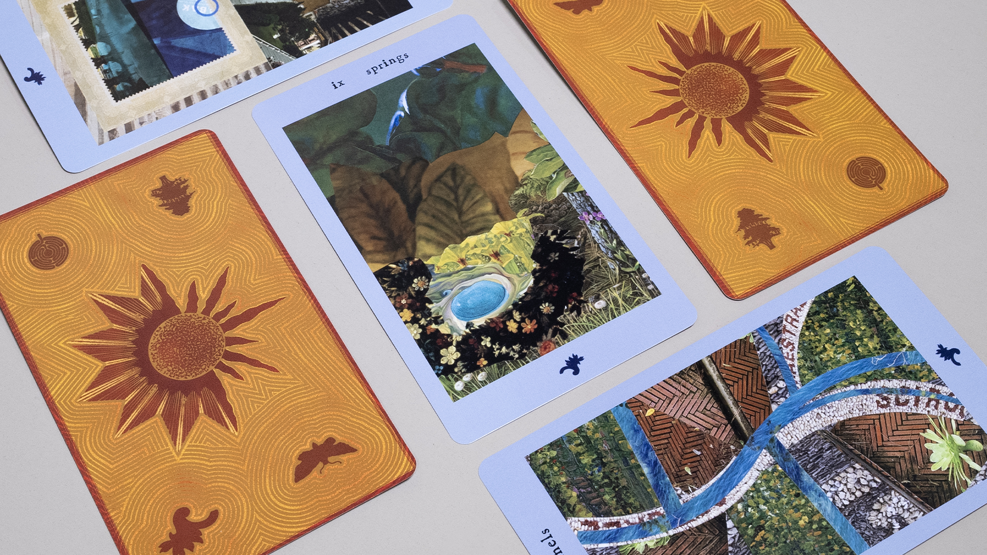 A tabletop arrangement of illustrated oracle-style cards featuring warm-toned sunburst backs and pastel blue-bordered fronts, including a central card labeled “ix springs” showing a collage of leaves, flowers, and a small blue pool, surrounded by other cards with nature-inspired imagery such as winding paths, water, foliage, and mosaic textures, photographed from an angled overhead view on a neutral surface.