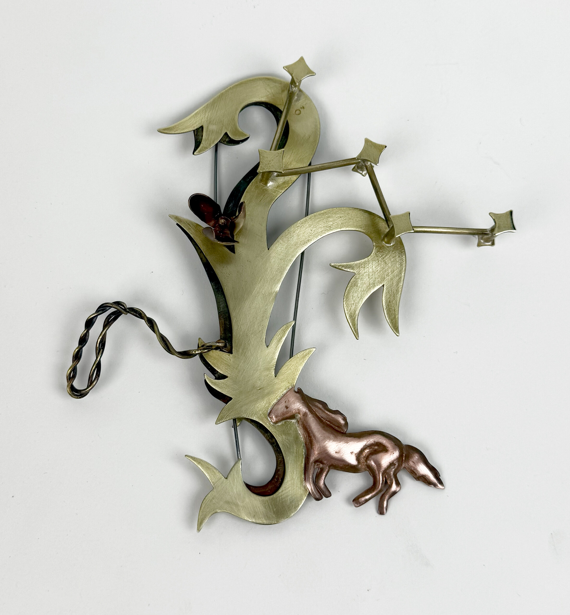 A decorative brooch featuring an abstract design made of metal, with curved, leafy shapes. The brooch includes a small, copper-colored horse figure attached to the design. It has a pin closure and some twisted metal details, suggesting a handmade appearance. The background is white, highlighting the brooch's intricate shapes and shiny finish.