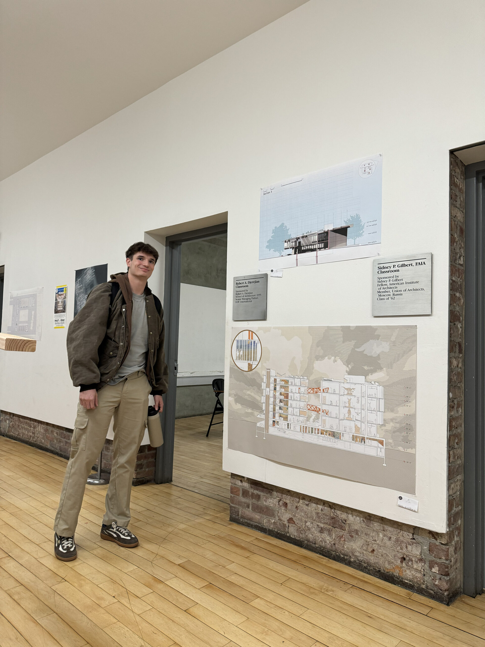 A young man stands in an interior space with wooden flooring, next to a large architectural illustration mounted on the wall. The illustration includes architectural sections and plans. Signage next to the artwork provides information about the exhibit. The man is wearing a brown jacket, light beige pants, and sneakers, holding a water bottle. In the background, there is additional architectural display material and a simple chair.