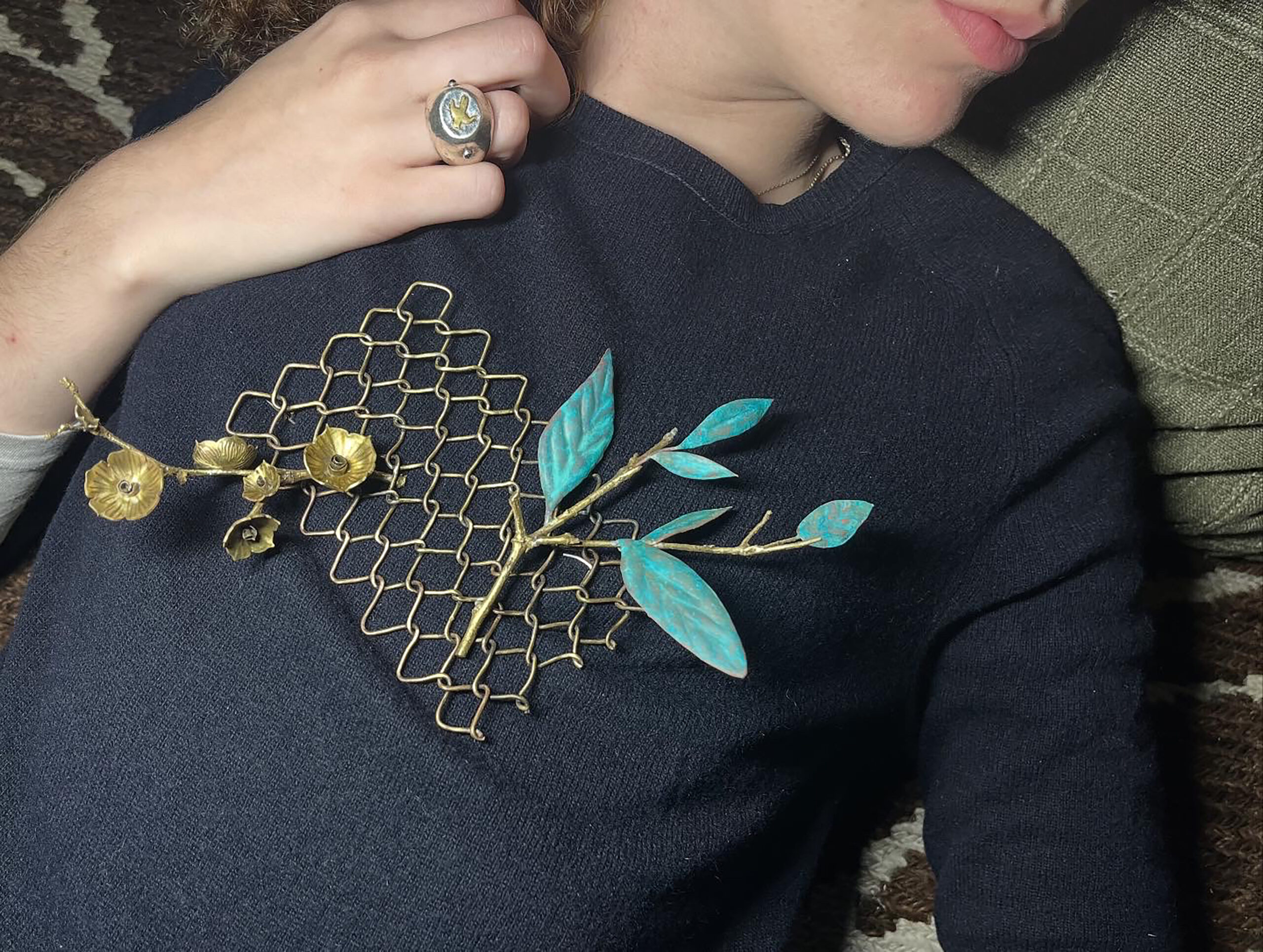A close-up image of a person reclining against a green pillow, wearing a dark sweater. A decorative brooch featuring metallic gold and turquoise leaves and flowers is attached to the sweater. The person's hand, adorned with a ring, rests near the brooch. The background consists of a patterned rug.