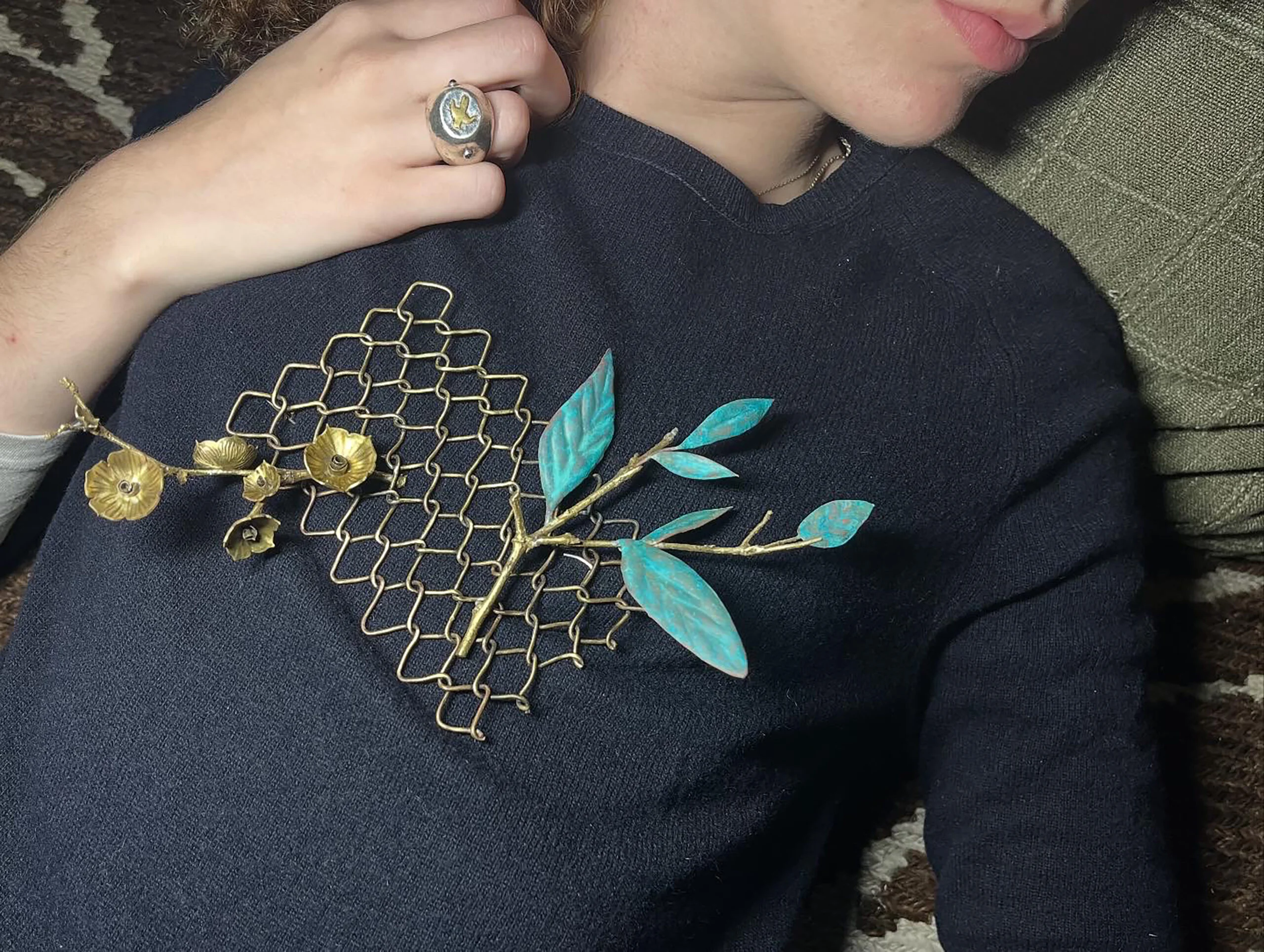 A close-up image of a person reclining against a green pillow, wearing a dark sweater. A decorative brooch featuring metallic gold and turquoise leaves and flowers is attached to the sweater. The person's hand, adorned with a ring, rests near the brooch. The background consists of a patterned rug.